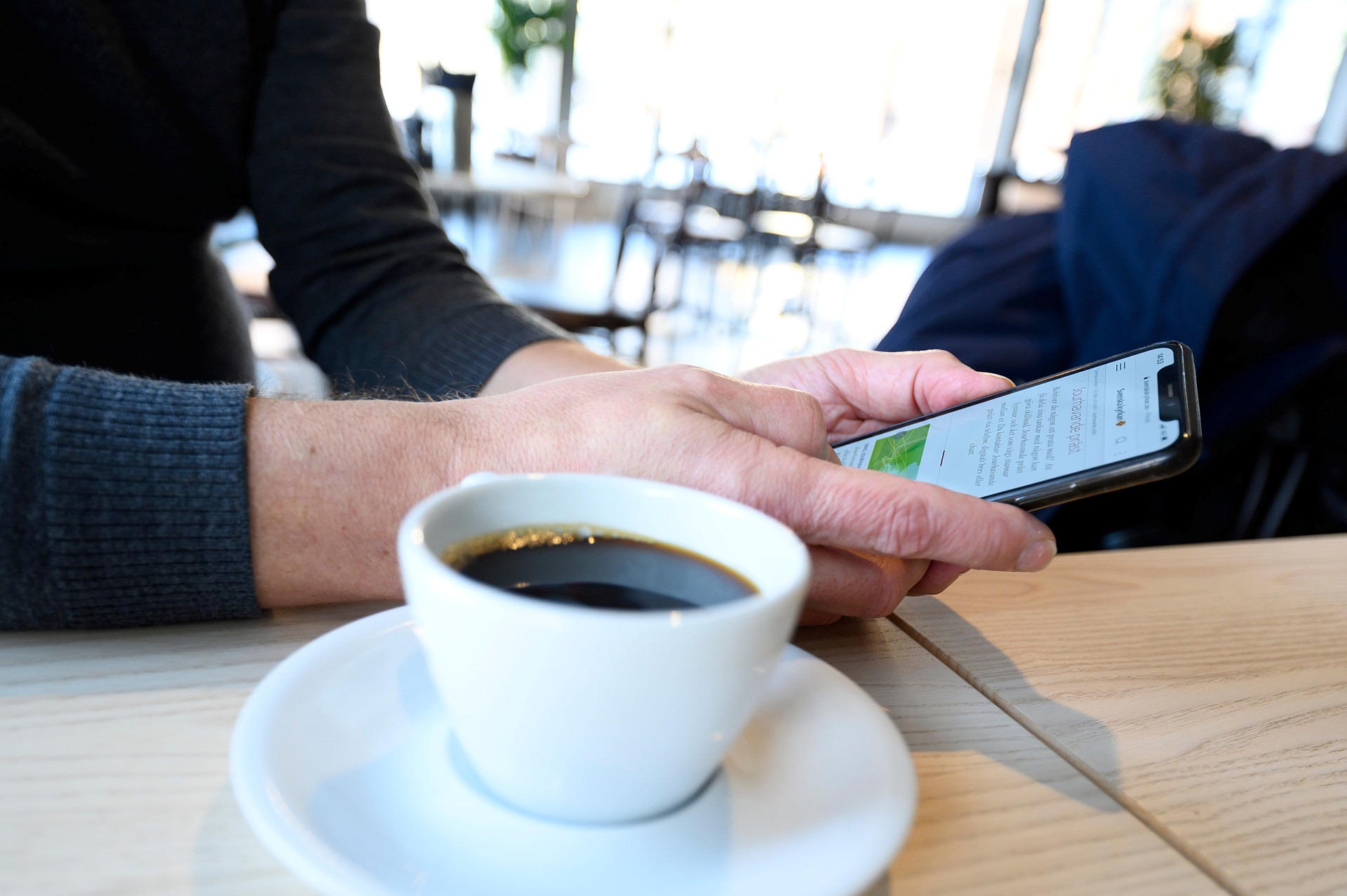 Kahvikuppi pöydälla ja käsi joka selaa kännykkää.