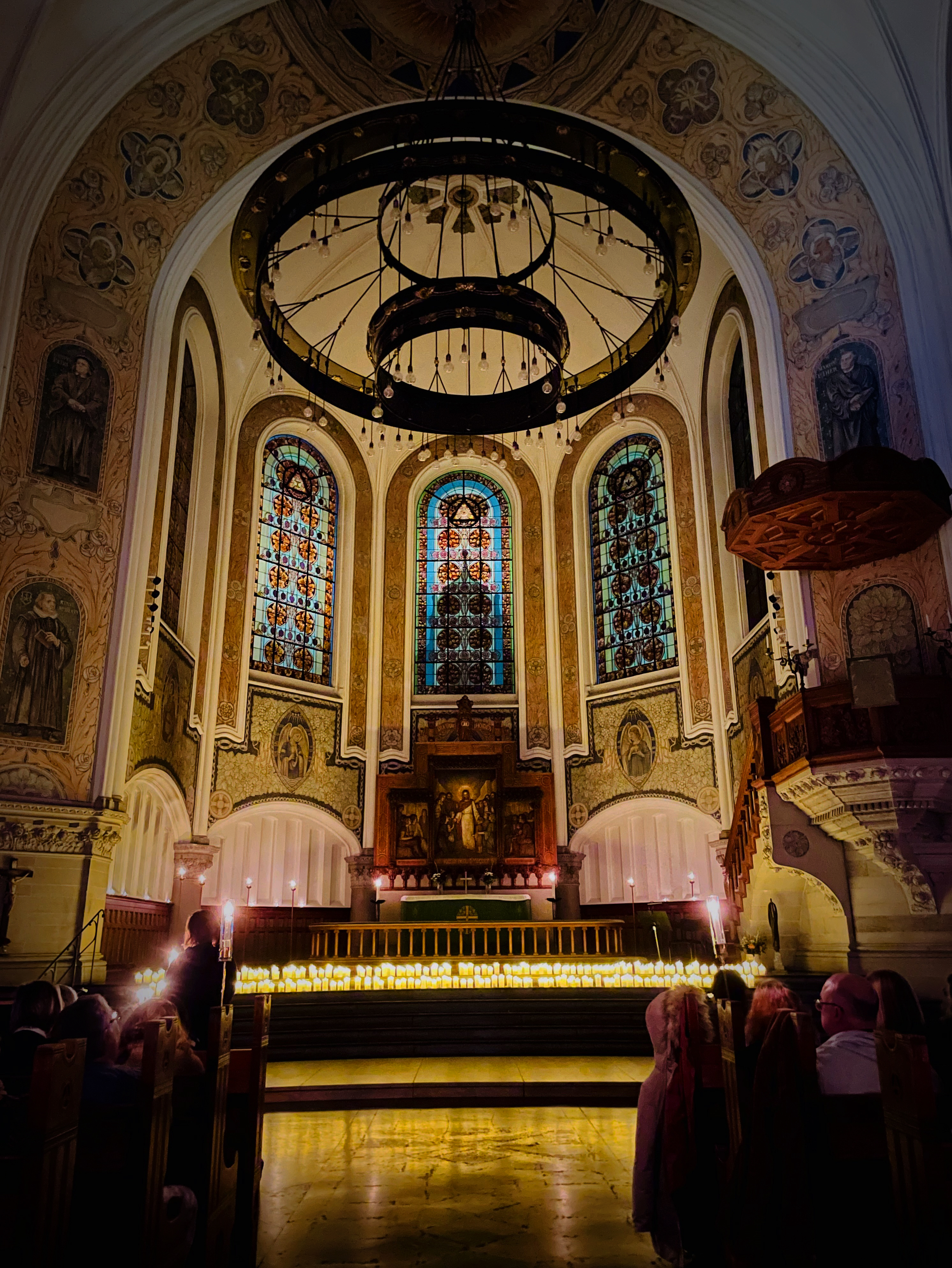 Kor i kyrka med tre stora fönster och altare. I trappan upp står hundratals tända ljus,