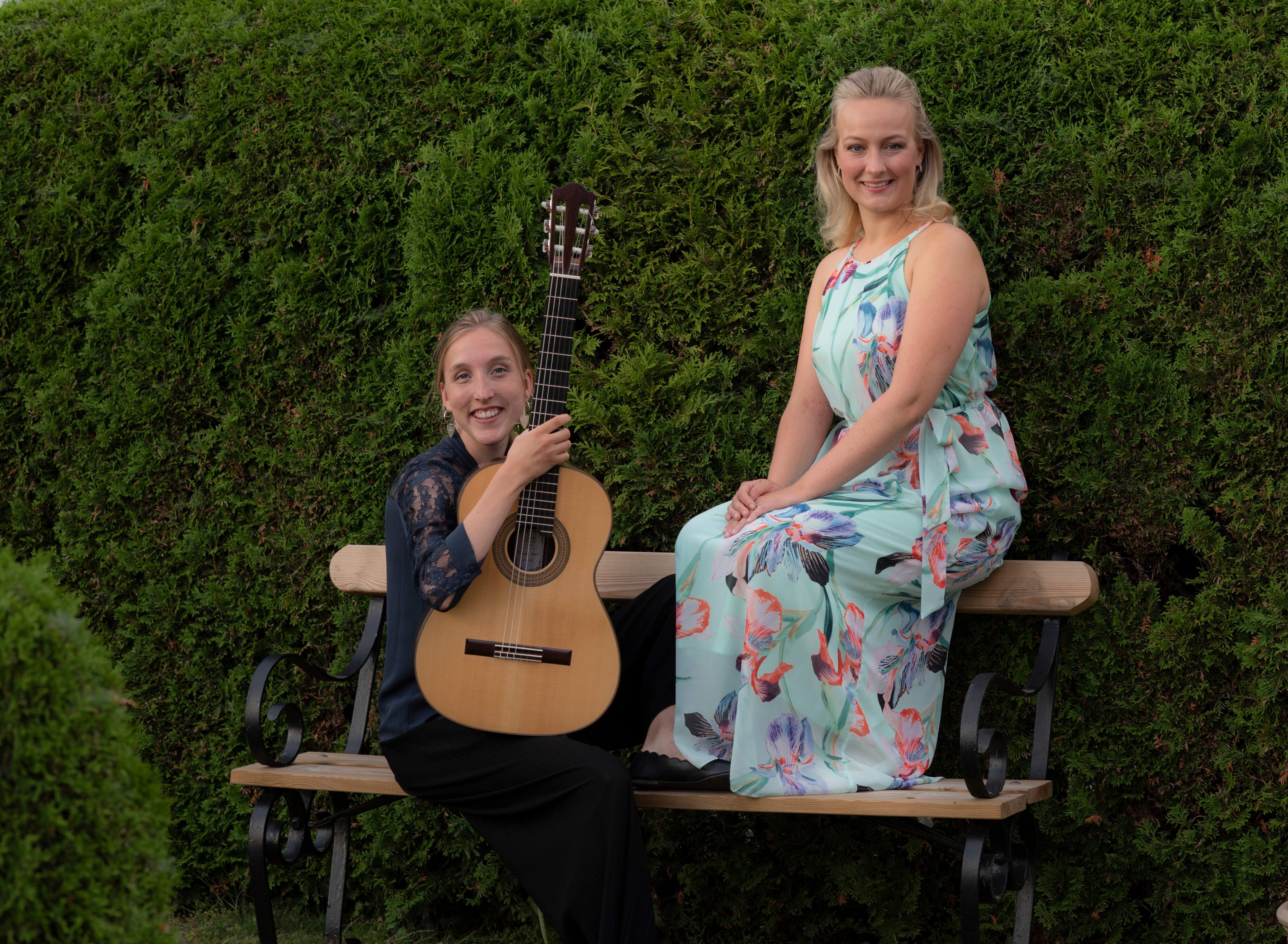 Två kvinnor sittande på en bänk, den ena med en gitarr i famnen. Gröna buskar i bakgrunden.