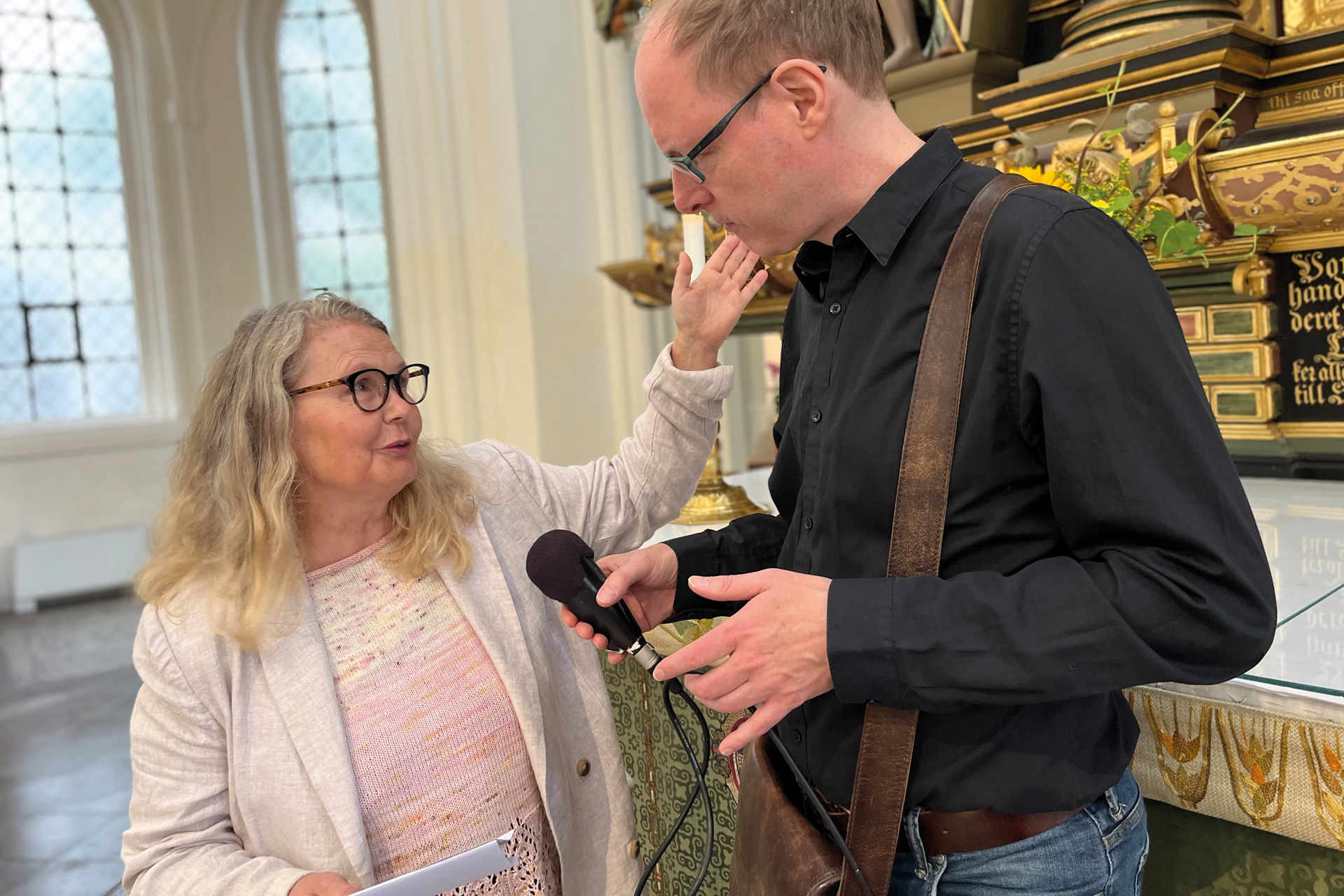 Kvinna med glasögon och blont långt vågigt hår i intervjusituation med en man med mikrofon i kyrkorum.