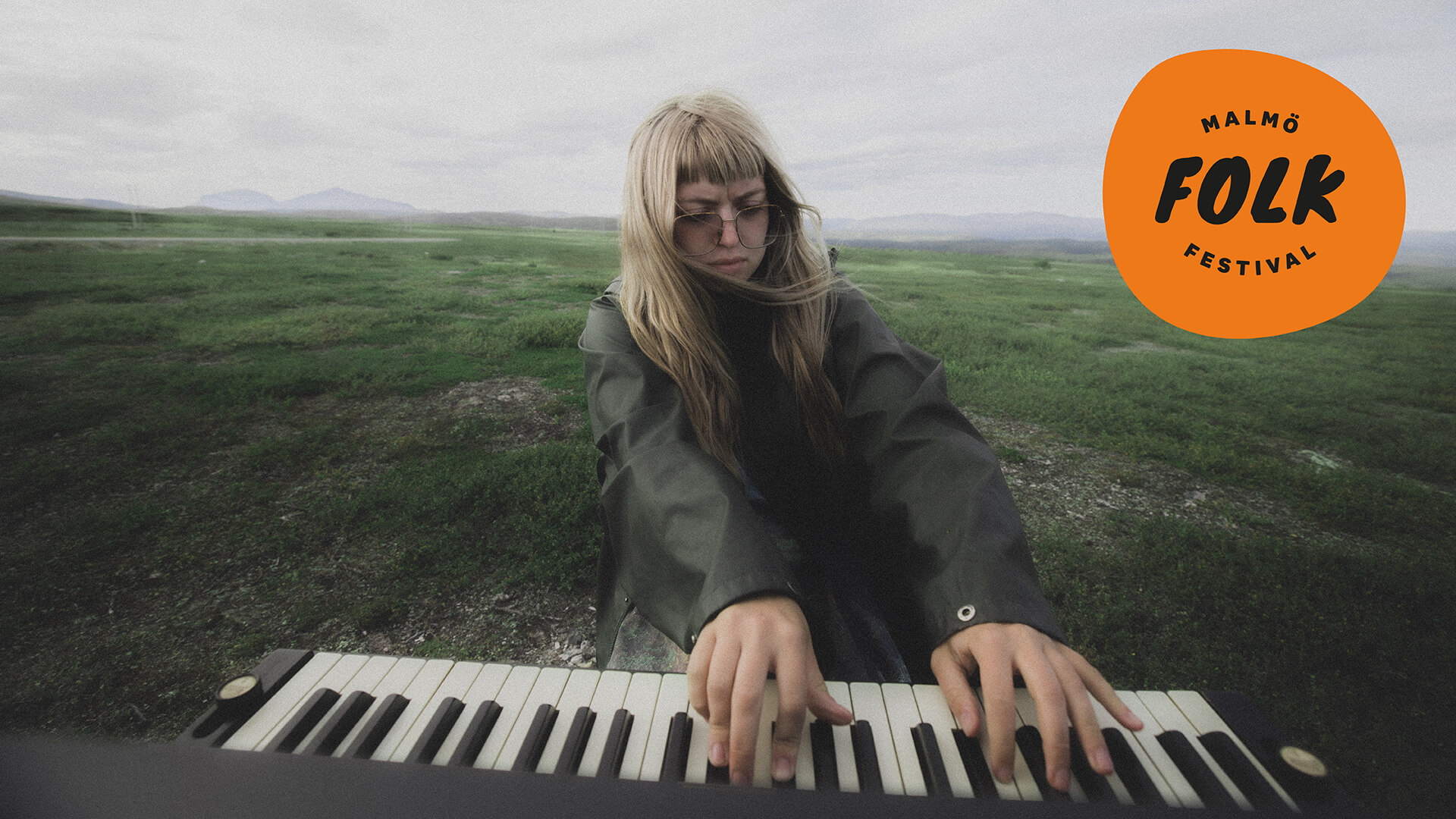 Bild av Sofia HK i ett kargt landskap och spelar på ett piano. På bilden finns en logga med texten Malmö folk festival.
