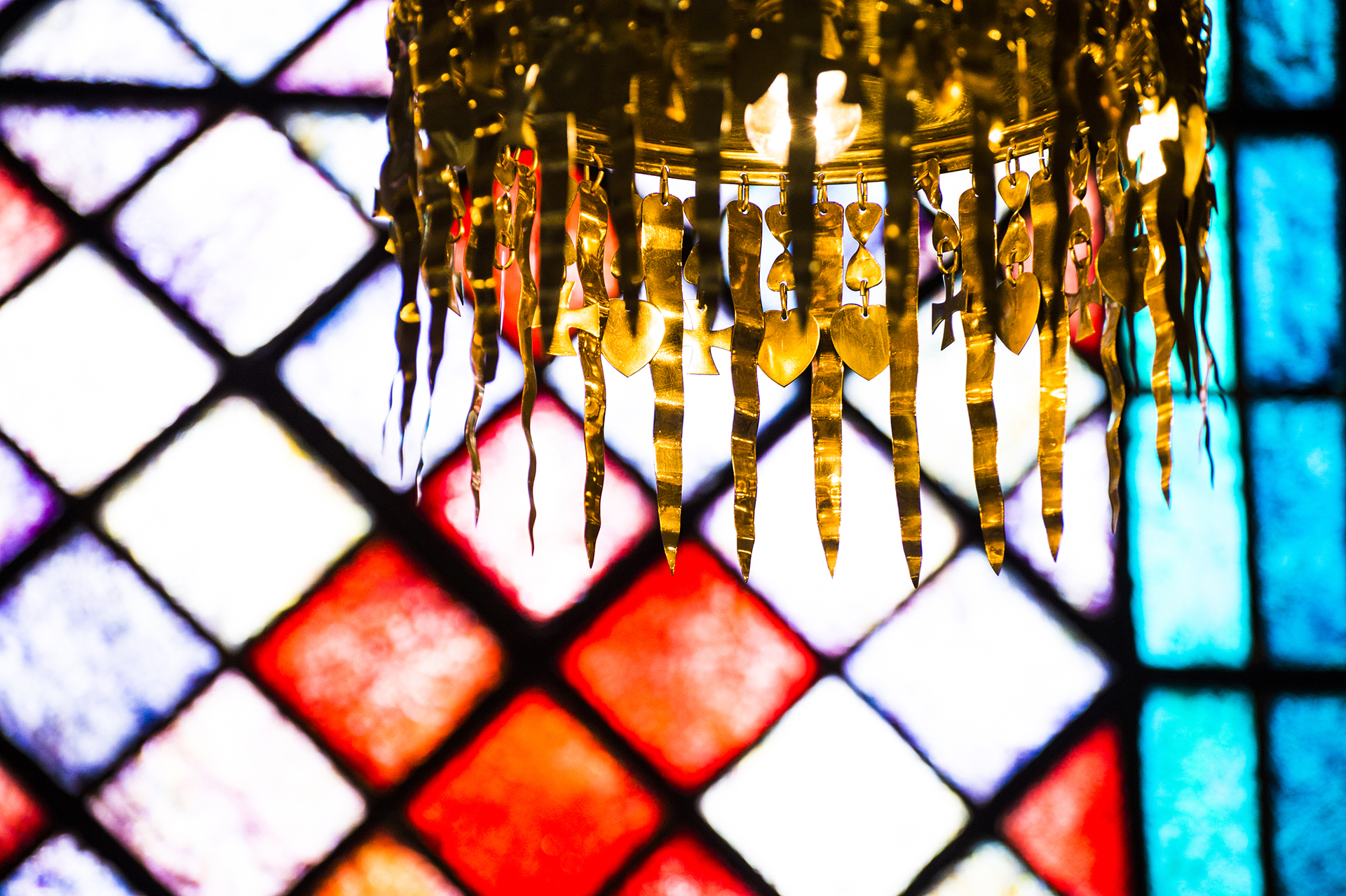 Ljuskrona i guld i förgrunden, i bakgrunden kyrkfönster i rött, lila och blått.