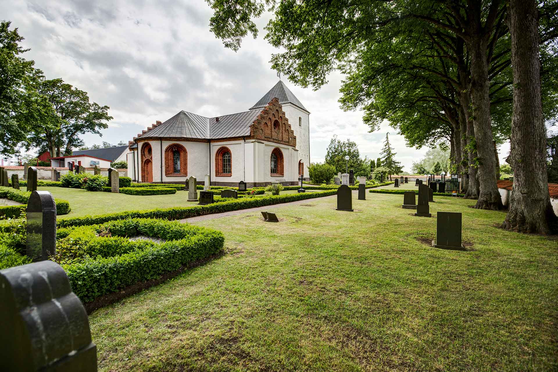 Vitputsad kyrka med röda tegeldetaljer omgiven av en lummig kyrkogård.