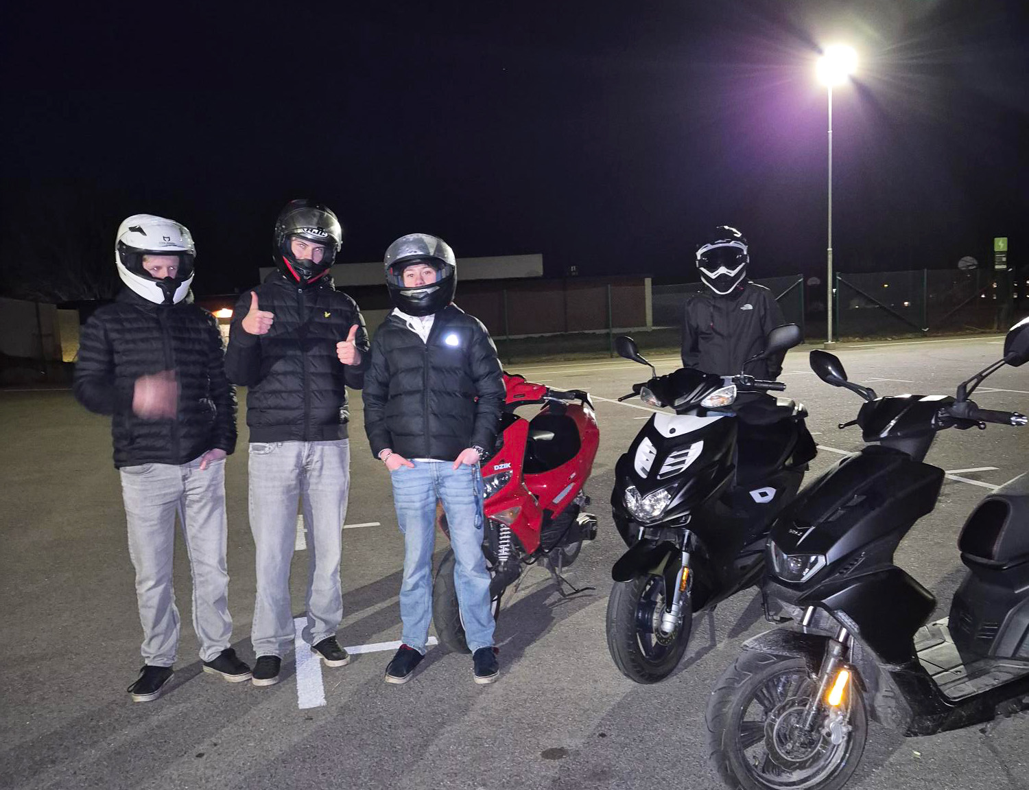 Tre tonårskillar med mopeder står på en parkering en mörk kväll.