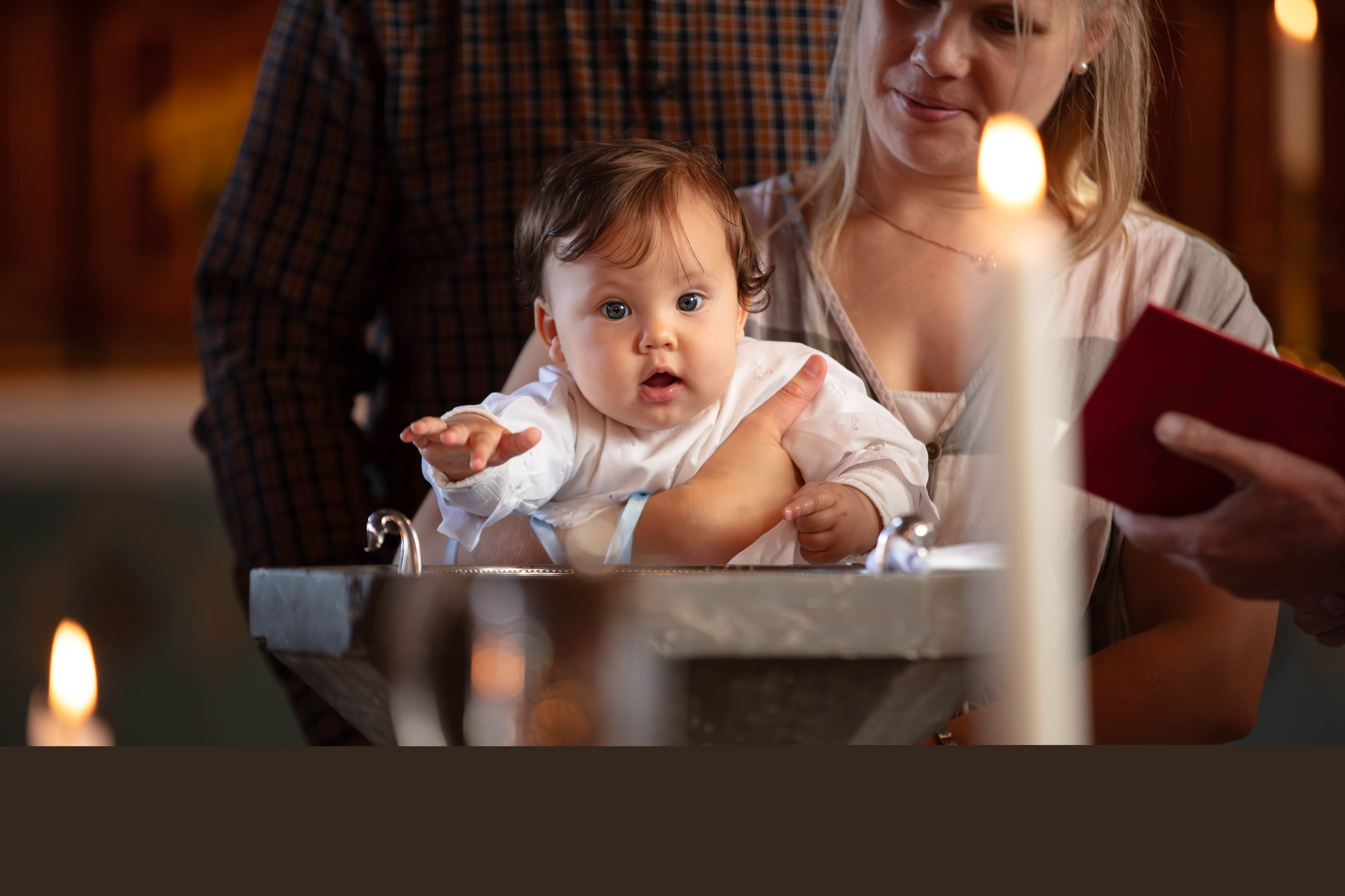 Dopbarn som hålls i mammans famn, vid dopfunten. I förgrunden står dopljuset tänt.