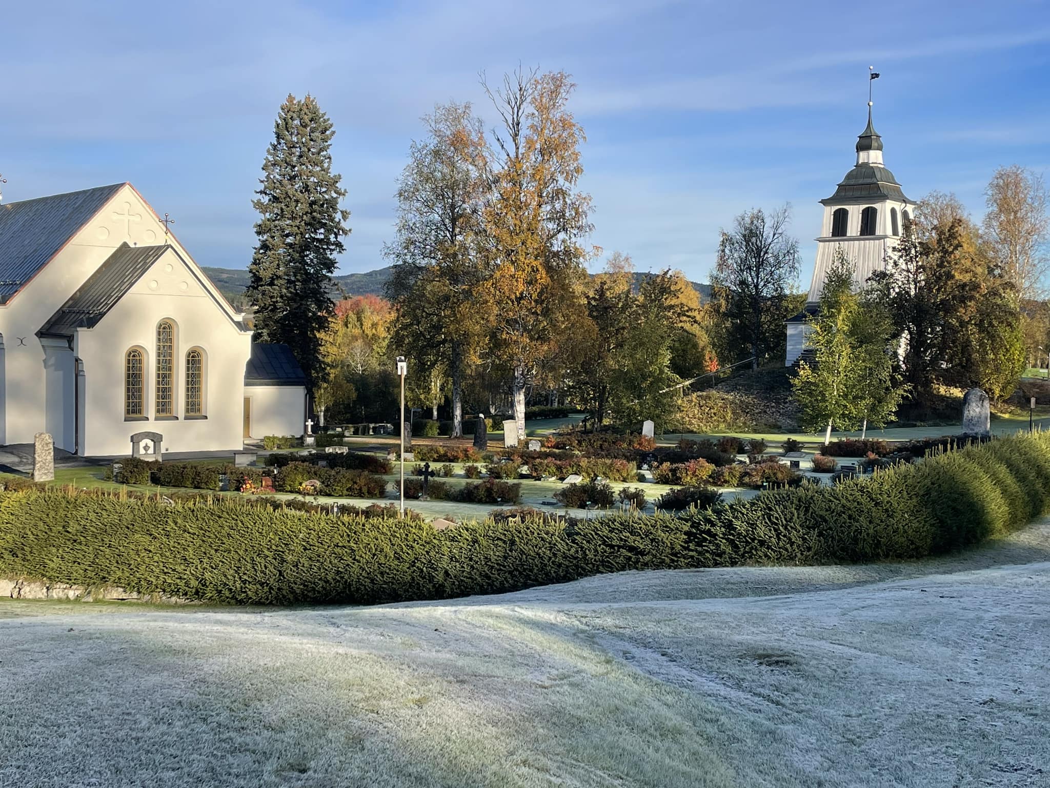 En bild av kyrka omgiven av frost belagda gräsytor och höstfärgade träd.