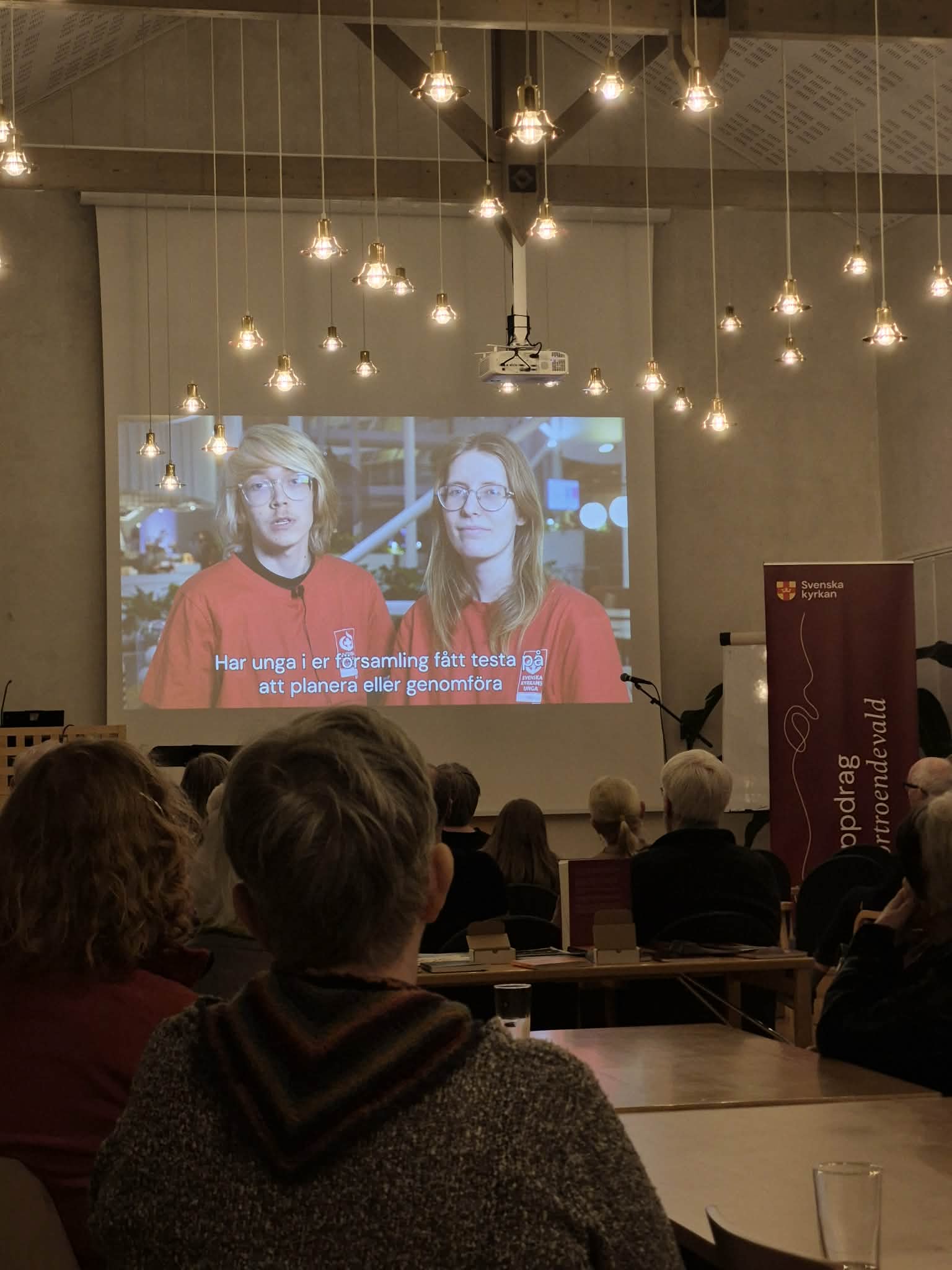 I en stor sal tittar många människor mot en projektorskärm där två unga personer pratar.