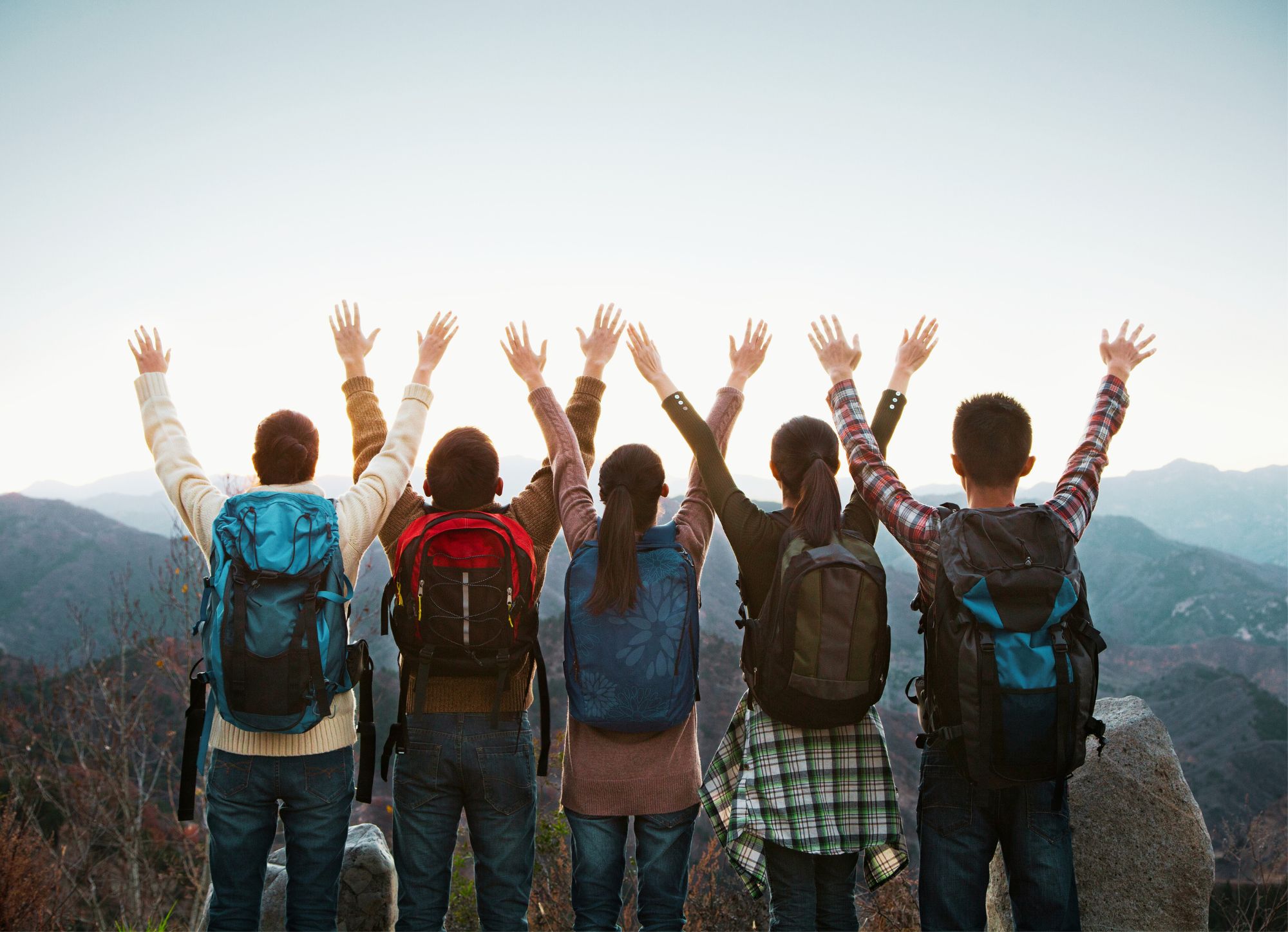 En grupp ungdomar med ryggsäckar står vänt mot berg och sträcker upp händerna i luften.