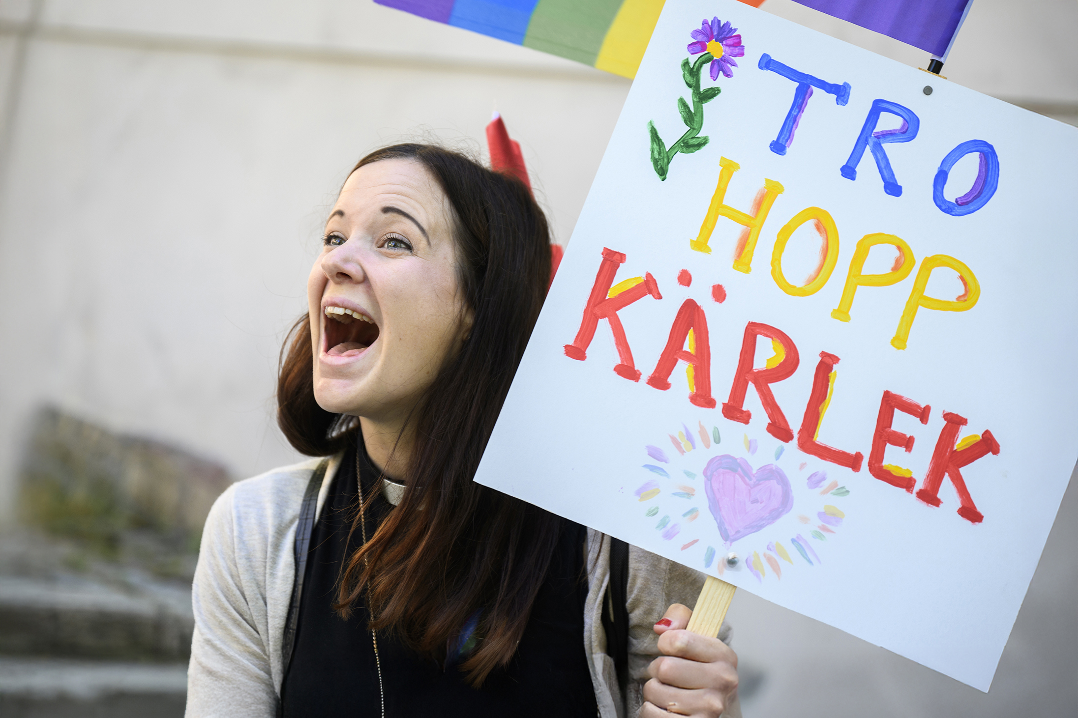 En kvinna med prästkrage som gapar och samtidigt håller ett plakat med texten tro, hopp, kärlek.