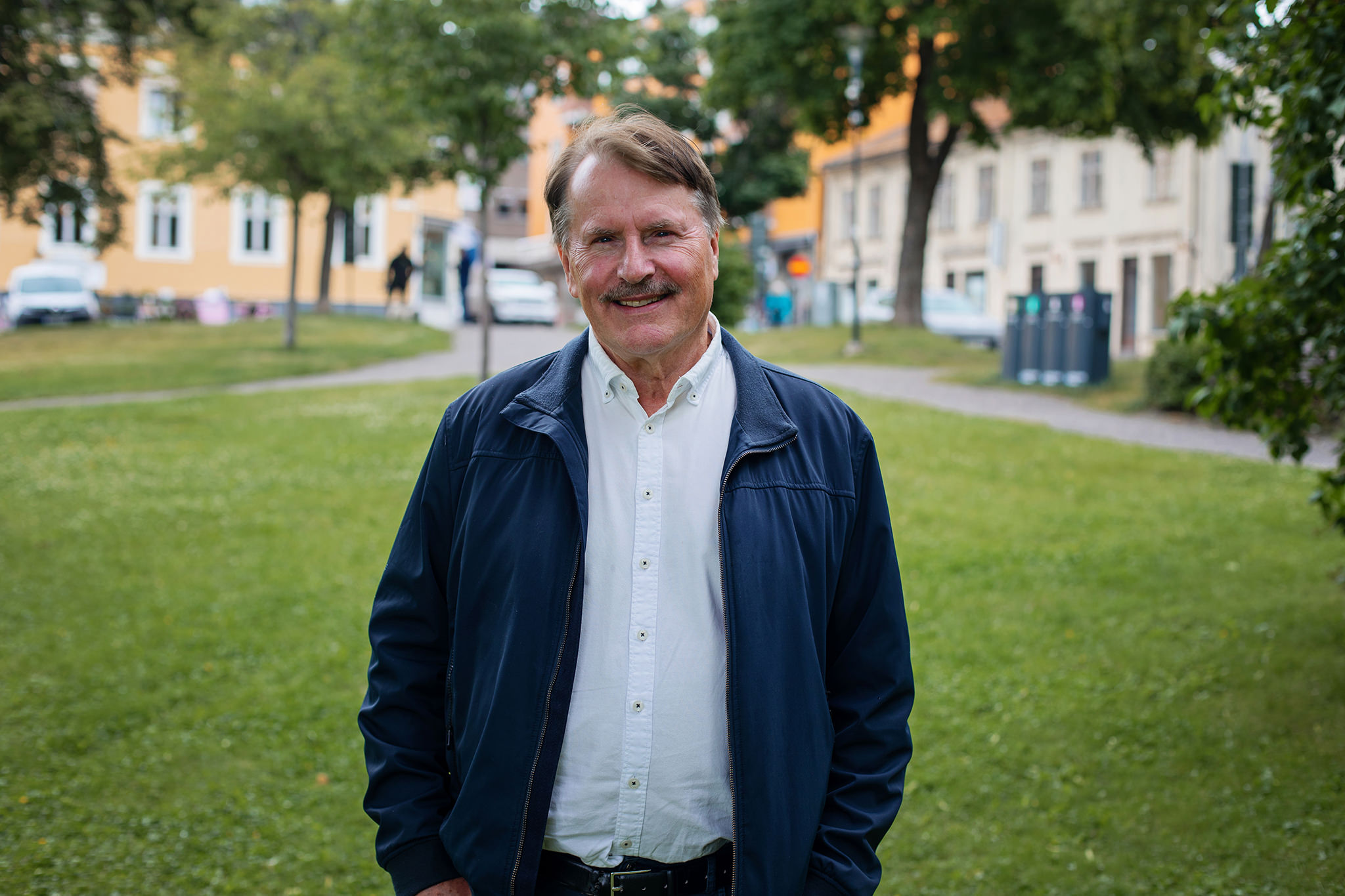 En man som står på en grön gräsmatta framför en gata med hus. Han är klädd i mörkblå jacka och vit skjorta.