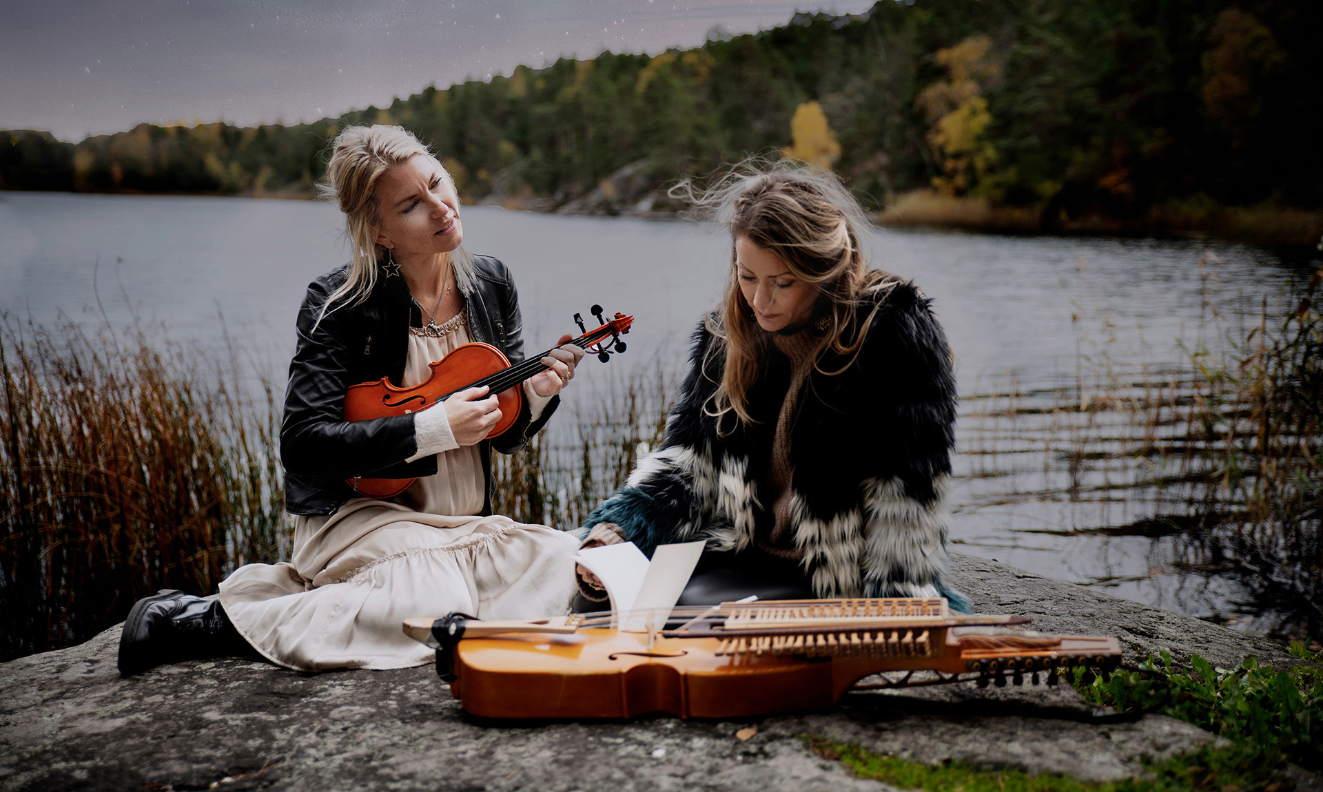 Folkmusikduon Erika och Cecila på en klippa vid en insjö kvällstid i dunkel sommarmiljö.