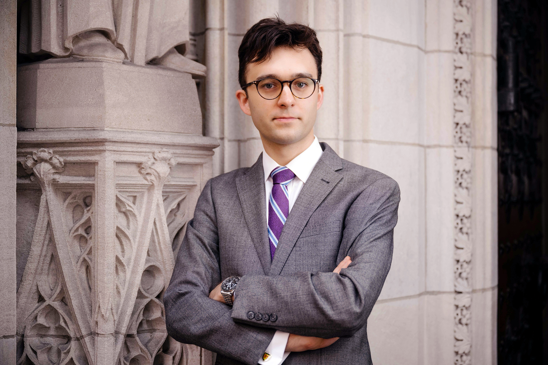 Nicolas Haigh, tillträdande organist i Uppsala domkyrka, portätterad i grå kostym, mörkt hår och glasögon mot bakgrund av en kyrkobyggnads exteriör.