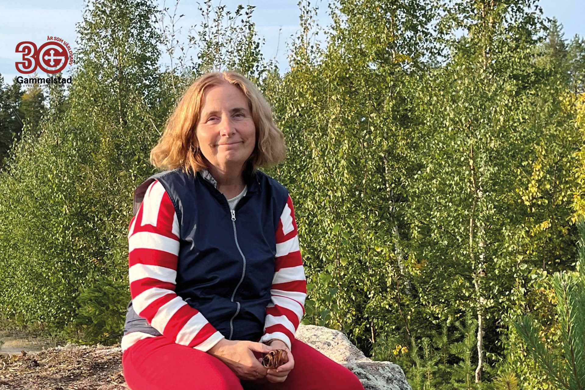 En kvinna sitter på en stenhäll med gröna björkar bakom sig. Hon har en blå väst, rödvitrandig tröja och röda byxor.