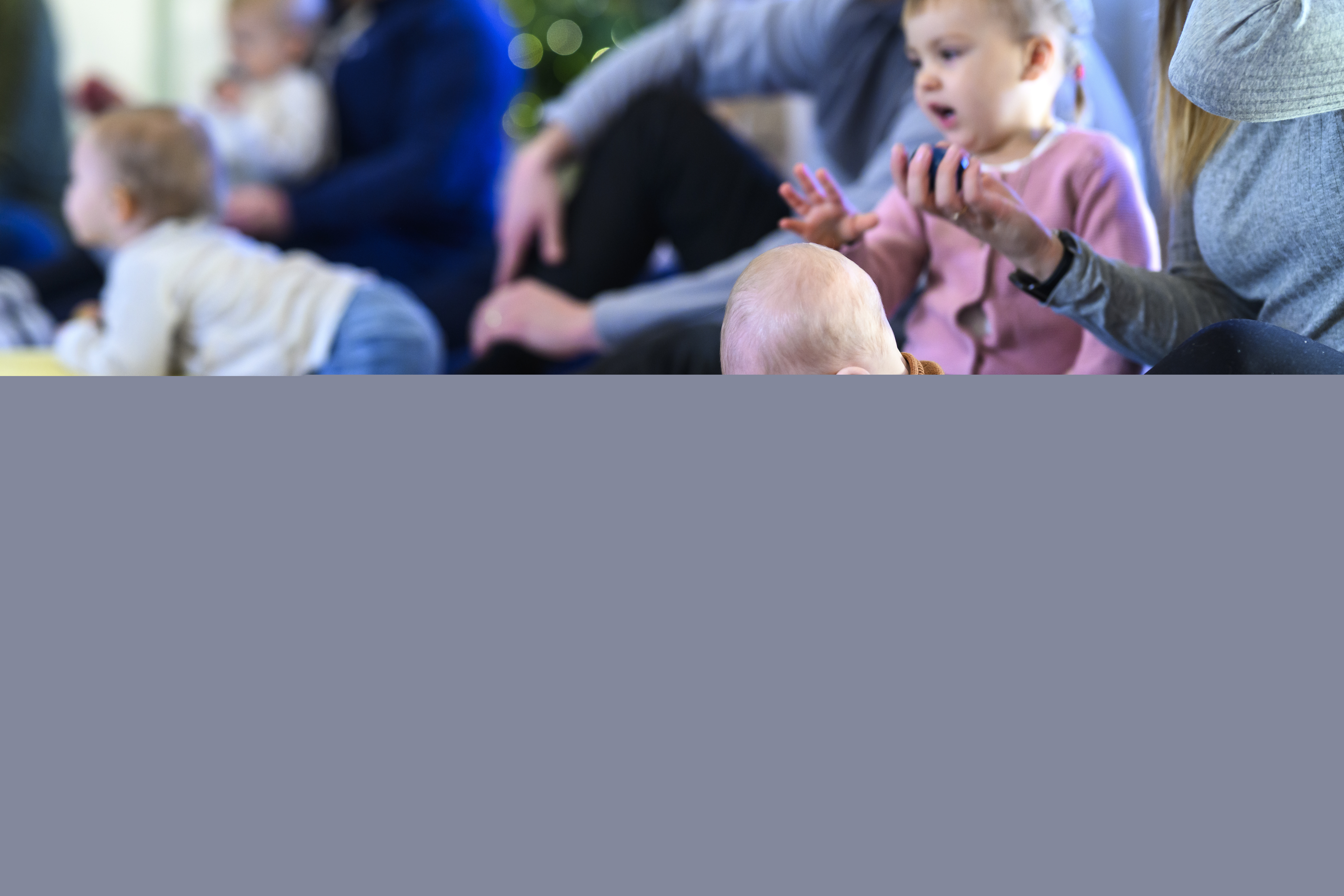 En baby sitter på golvet och leker meden boll. Runt omkring ser man syskon och föräldrar som sjunger.