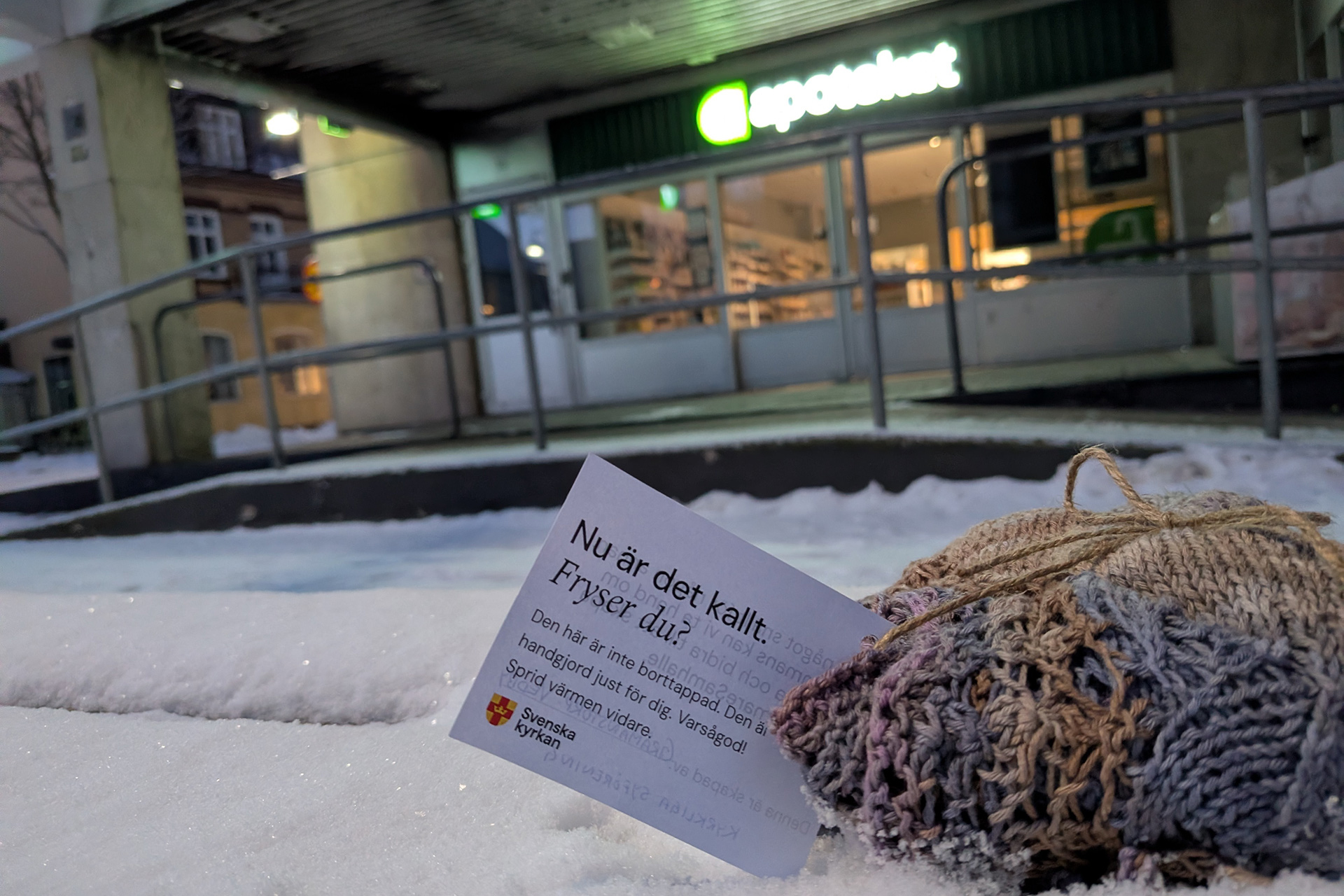 Ett stickat plagg på en snöig bänk framför ett apotek.