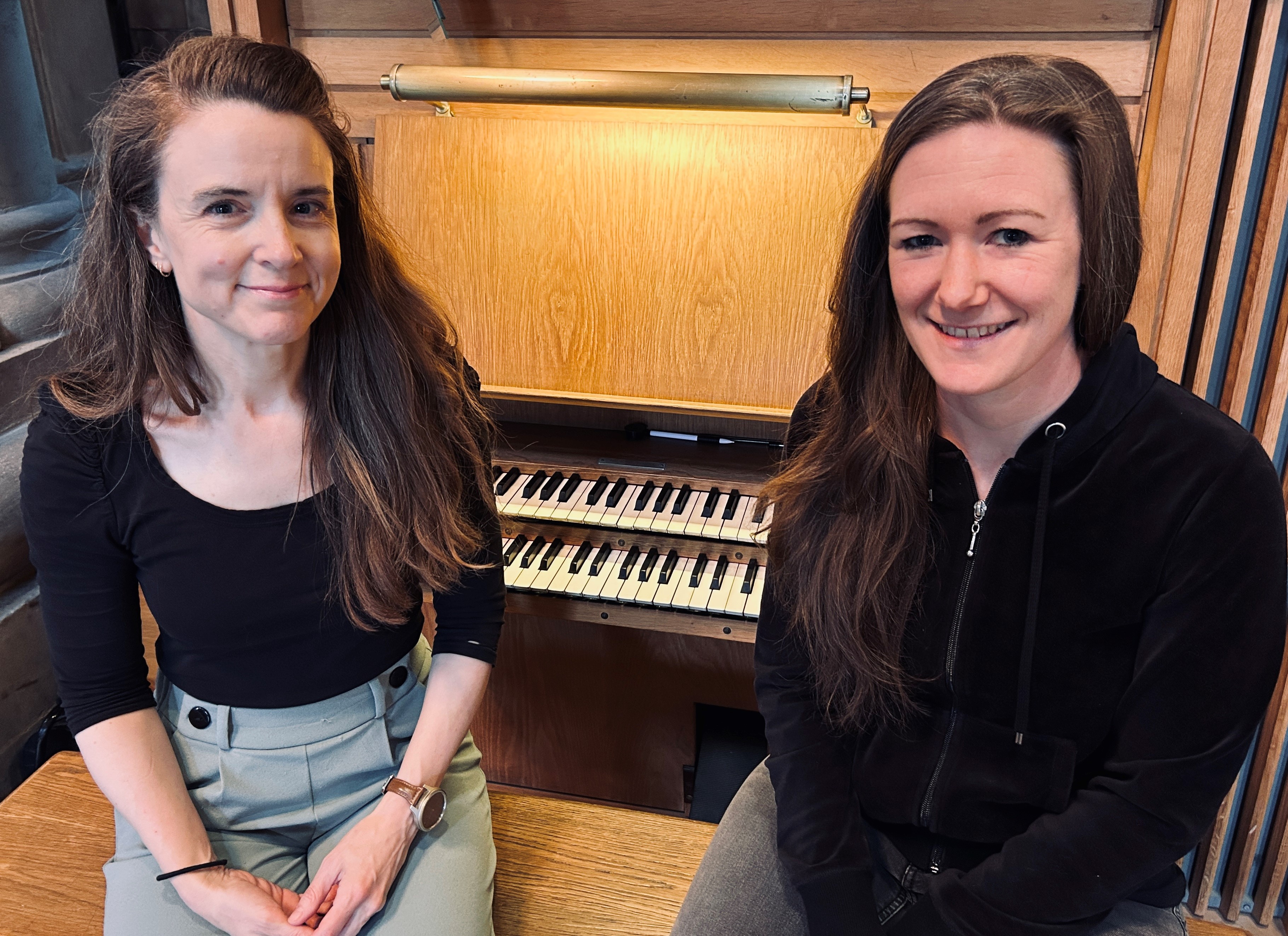 Två kvinnor med långt mörkt hår sitter på en pall framför en orgel.