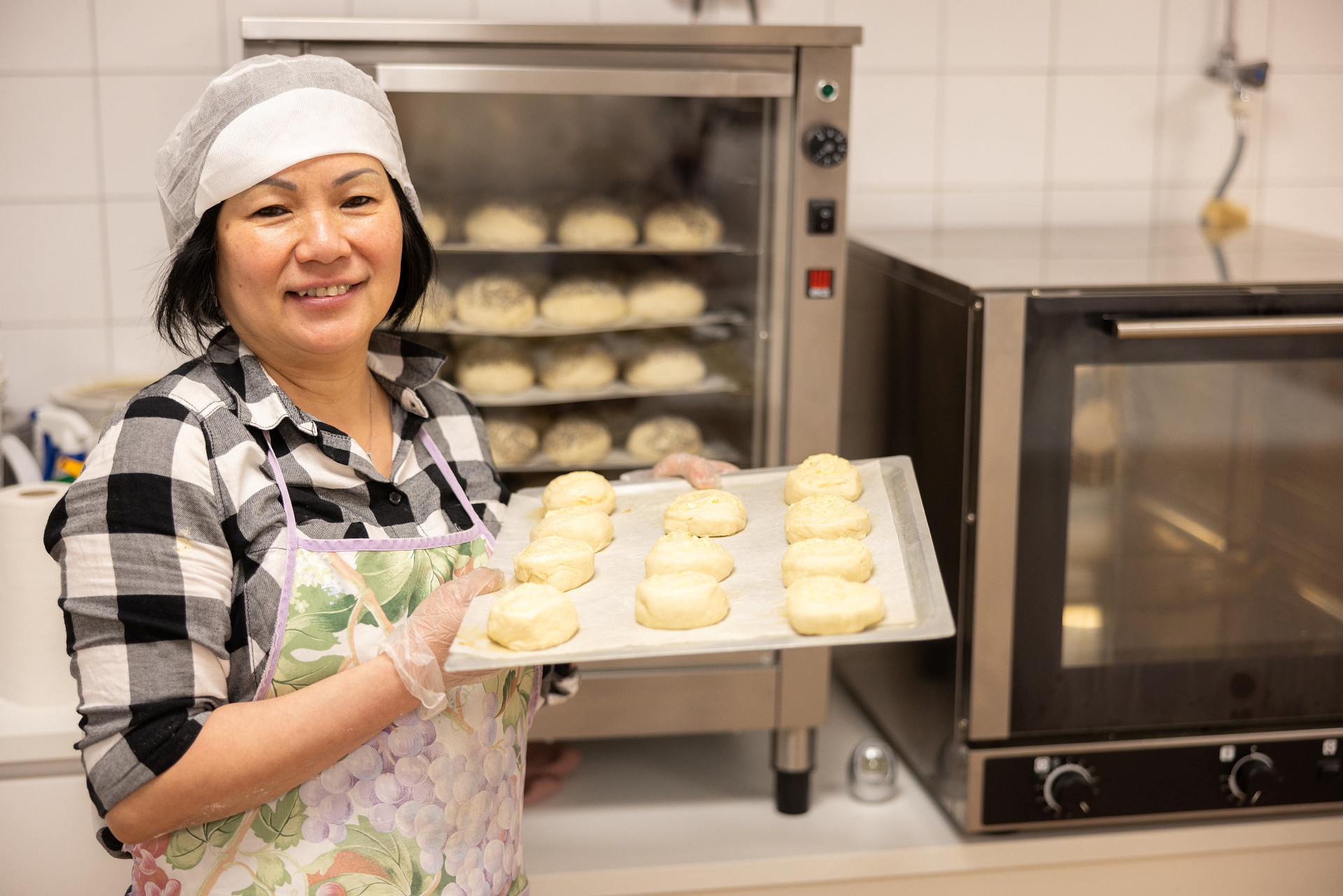 En kvinna står framför en stor ugn och visar stolt upp en bakplåt med frallor.
