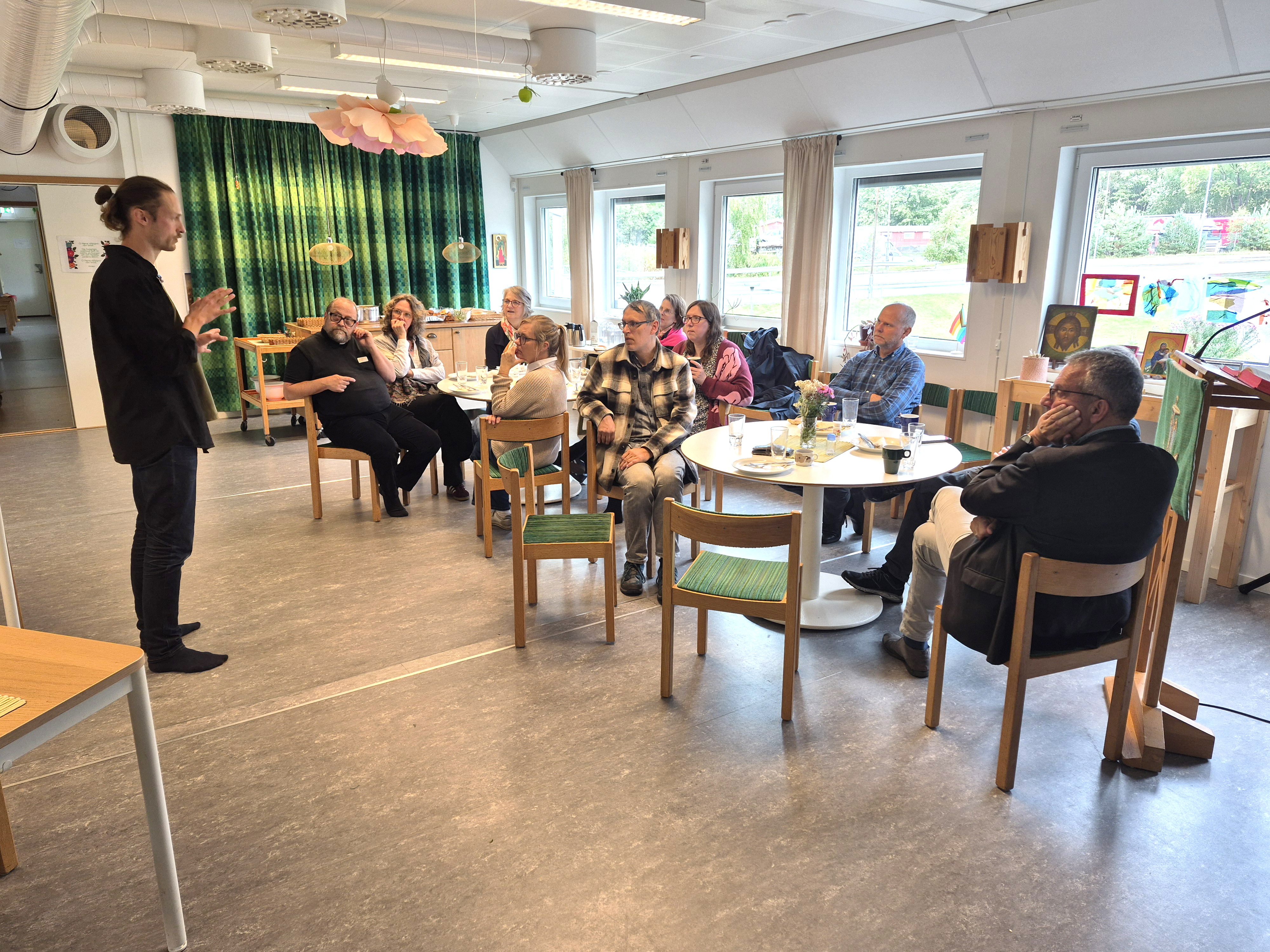 I en församlingssal står en person och berättar något för en grupp som sitter och lyssnar.