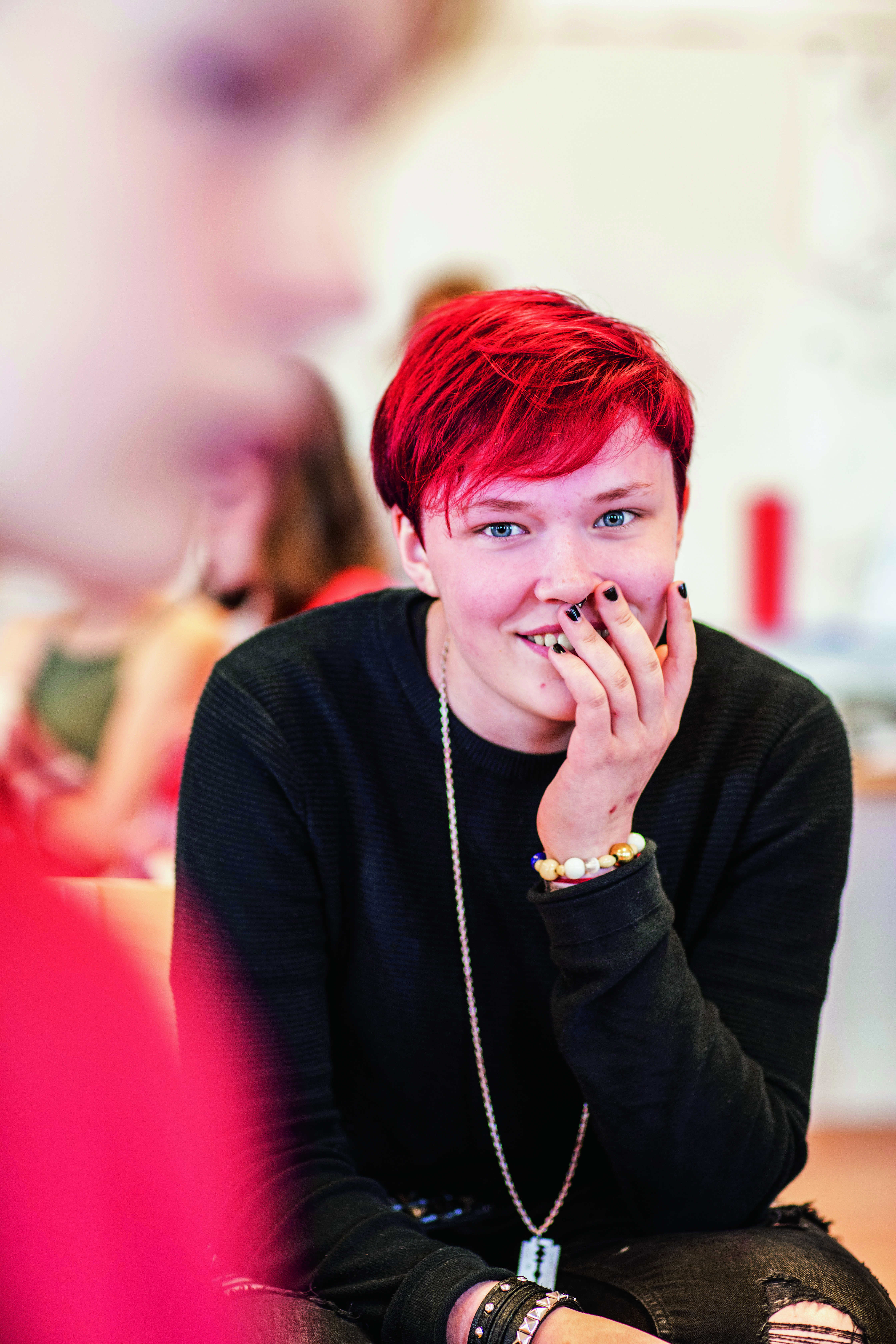 Ung person med rött, kort hår och svartmålade naglar vilar sin hand över munnen och verkar se nyfiket på något.