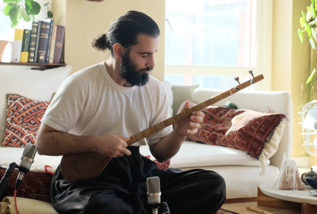 Musikern Koosha Atashgard, i vit t-shirt och med mörkt skägg,  sitter i ett ljust upplyst rum och spelar på stränginstrumentet santoor.