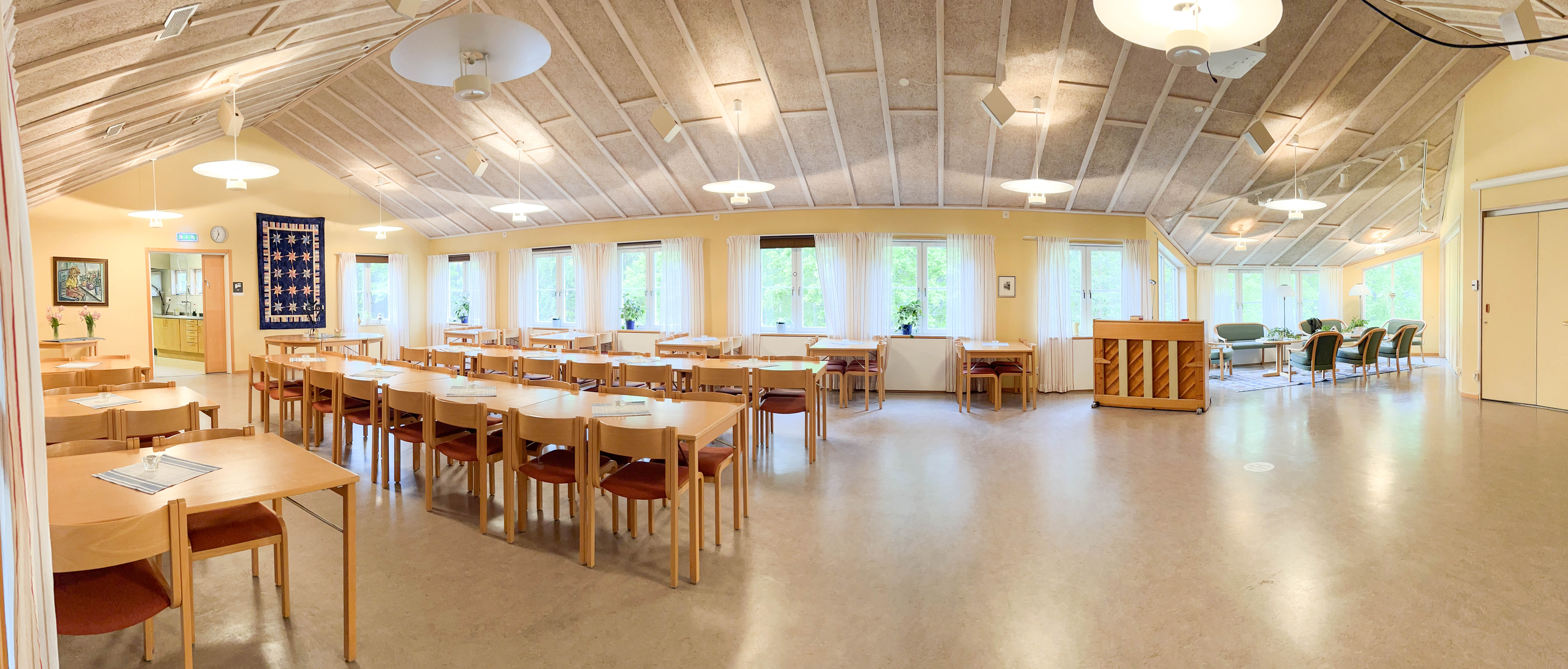 Panoramabild över församlingssalen. Till vänster står långbord och bord uppradade, till vänster är golvytan öppen. Bakom står ett piano och en soffgrupp utställt.