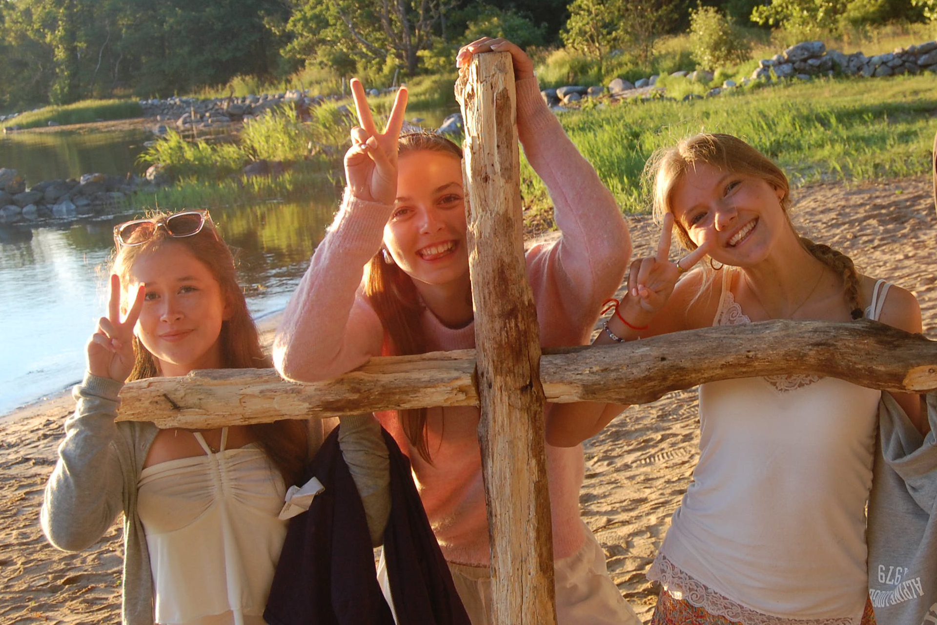 Tre glada tjejer står på en strand och håller ett träkors en vacker sommarkväll.