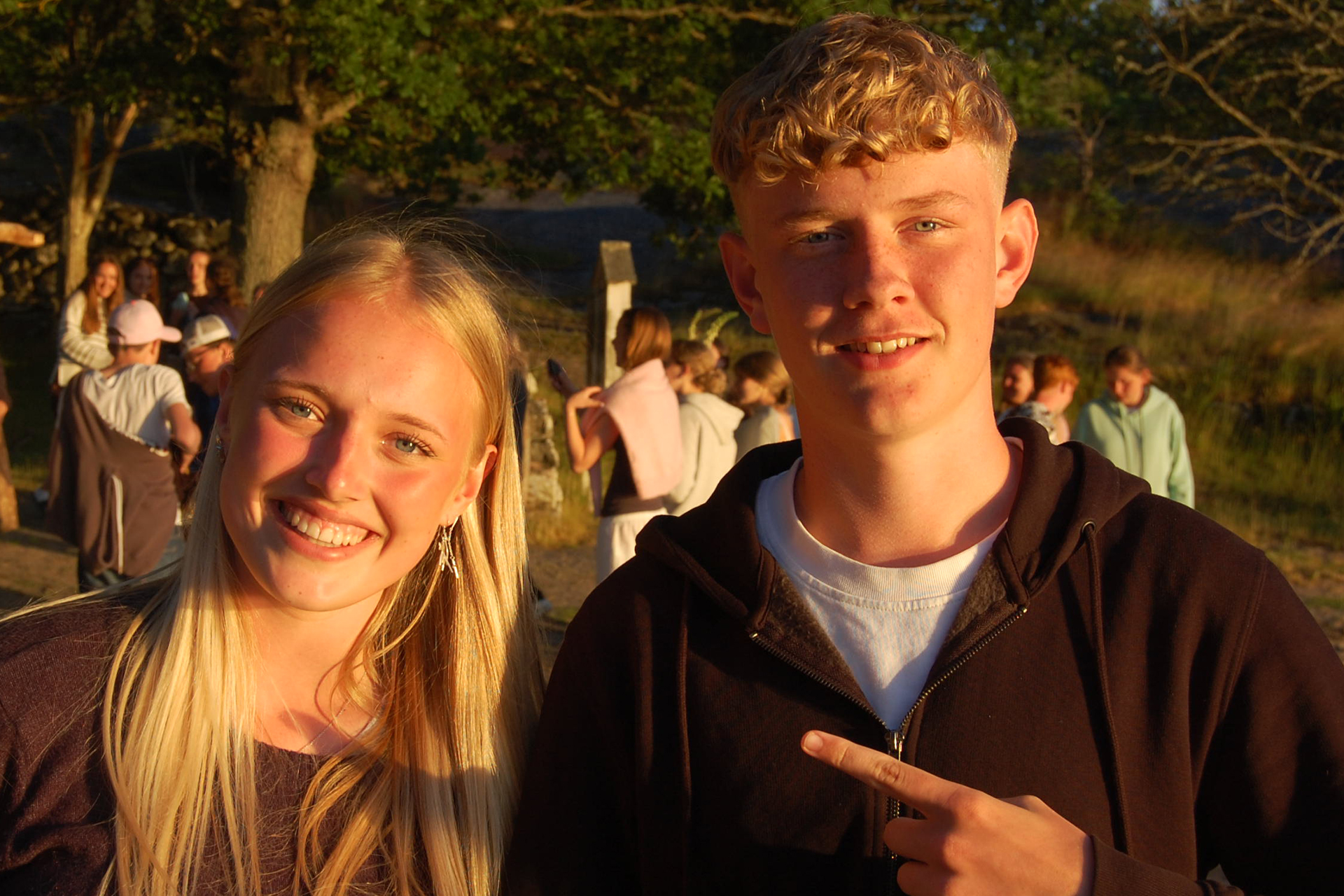 Två glada ungdomar står på en naturskön plats i sensommarsolen. I bakgrunen ses fler ungdomar och träd. 