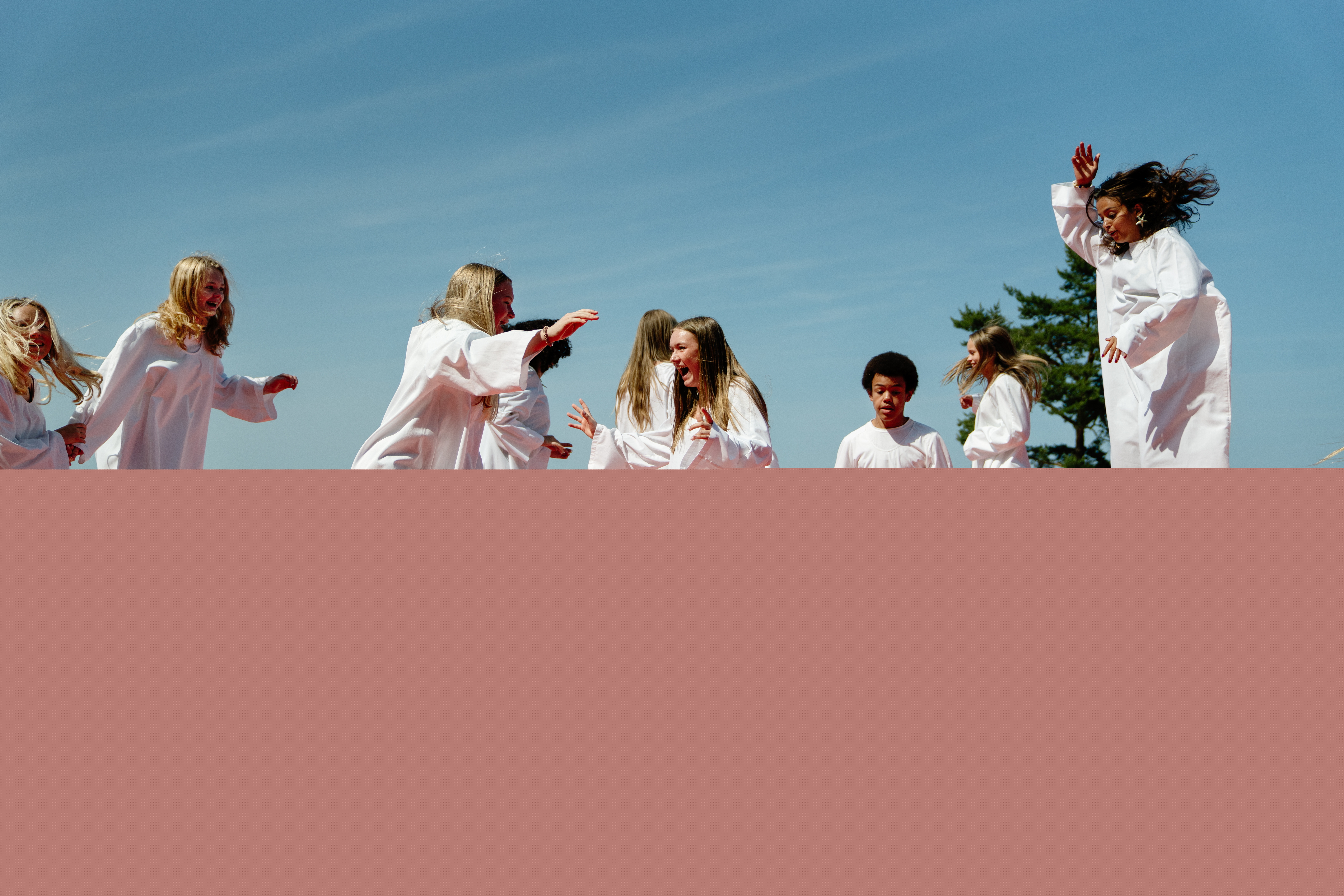Åtta konfirmander klädda i vita kåpor hoppar på en studsig madrass på en strand under sommartid. De har väldigt roligt.