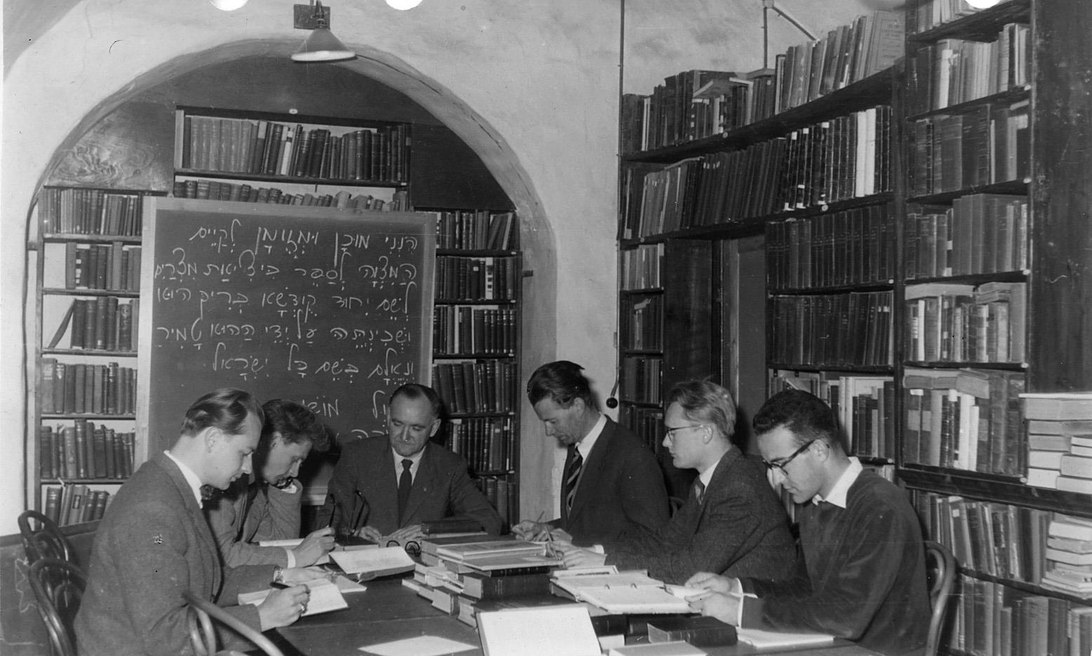 Studenter sitter kring ett bord med uppslagna böcker. Bokhyllor i bakgrunden.