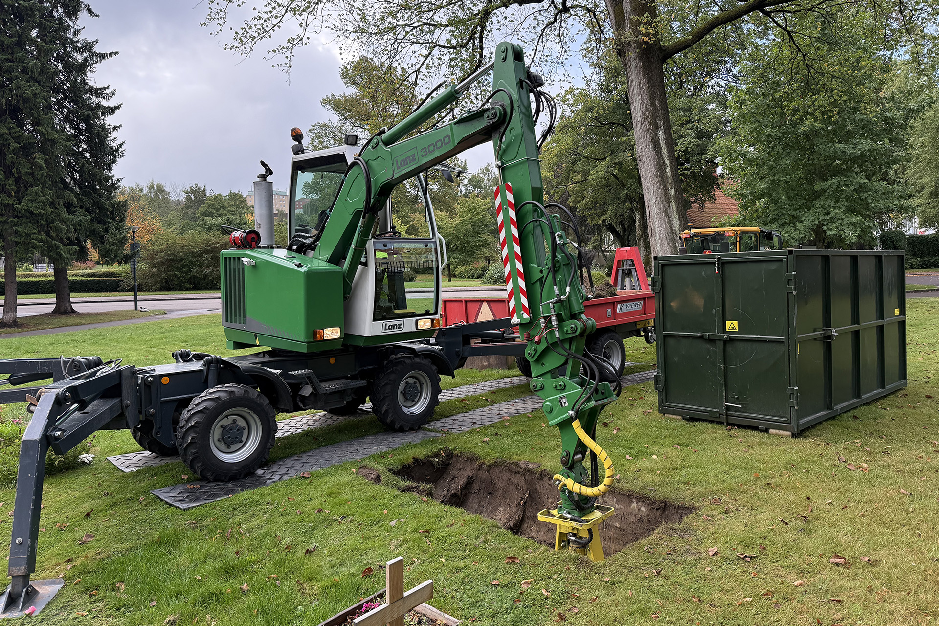Grävmaskin med skopa gräver en grav på en kyrkogård. I bakgrunden en grön container där jordmassorna släpps ned.