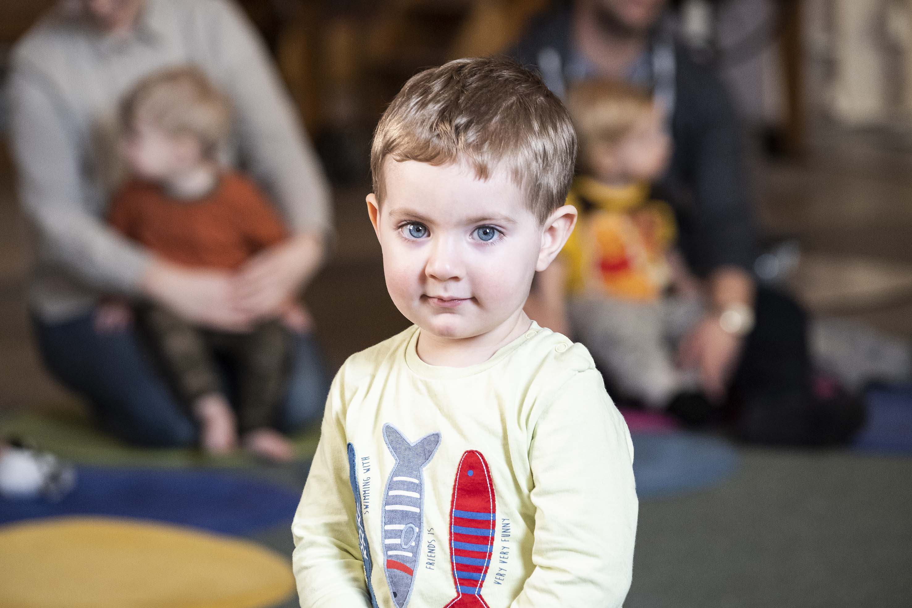 Pojke med fiskar på en tröja står framför vuxna som sitter på golvet i en kyrka. 