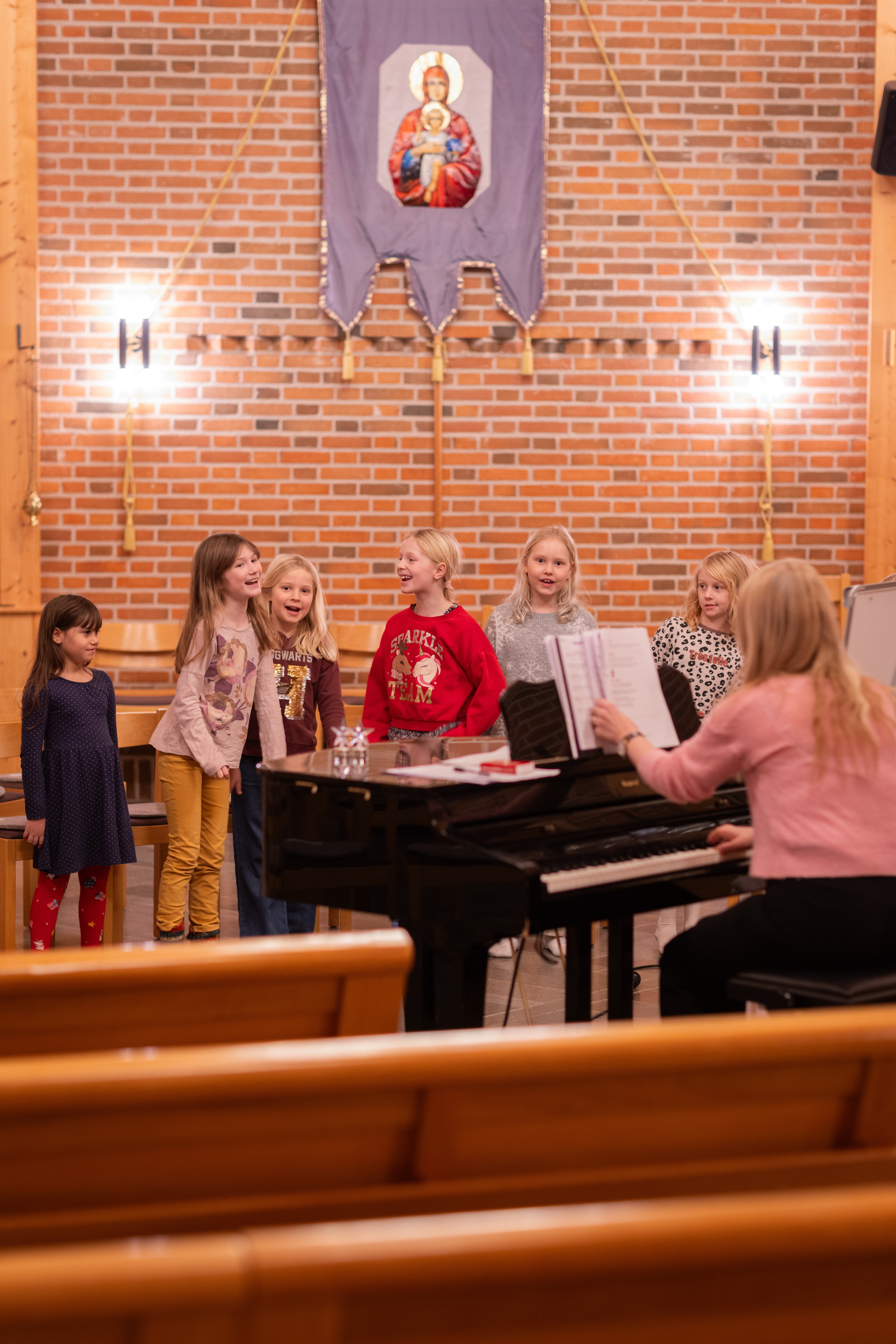 En grupp barn står runt en flygel i en kyrka och skrattar och sjunger.