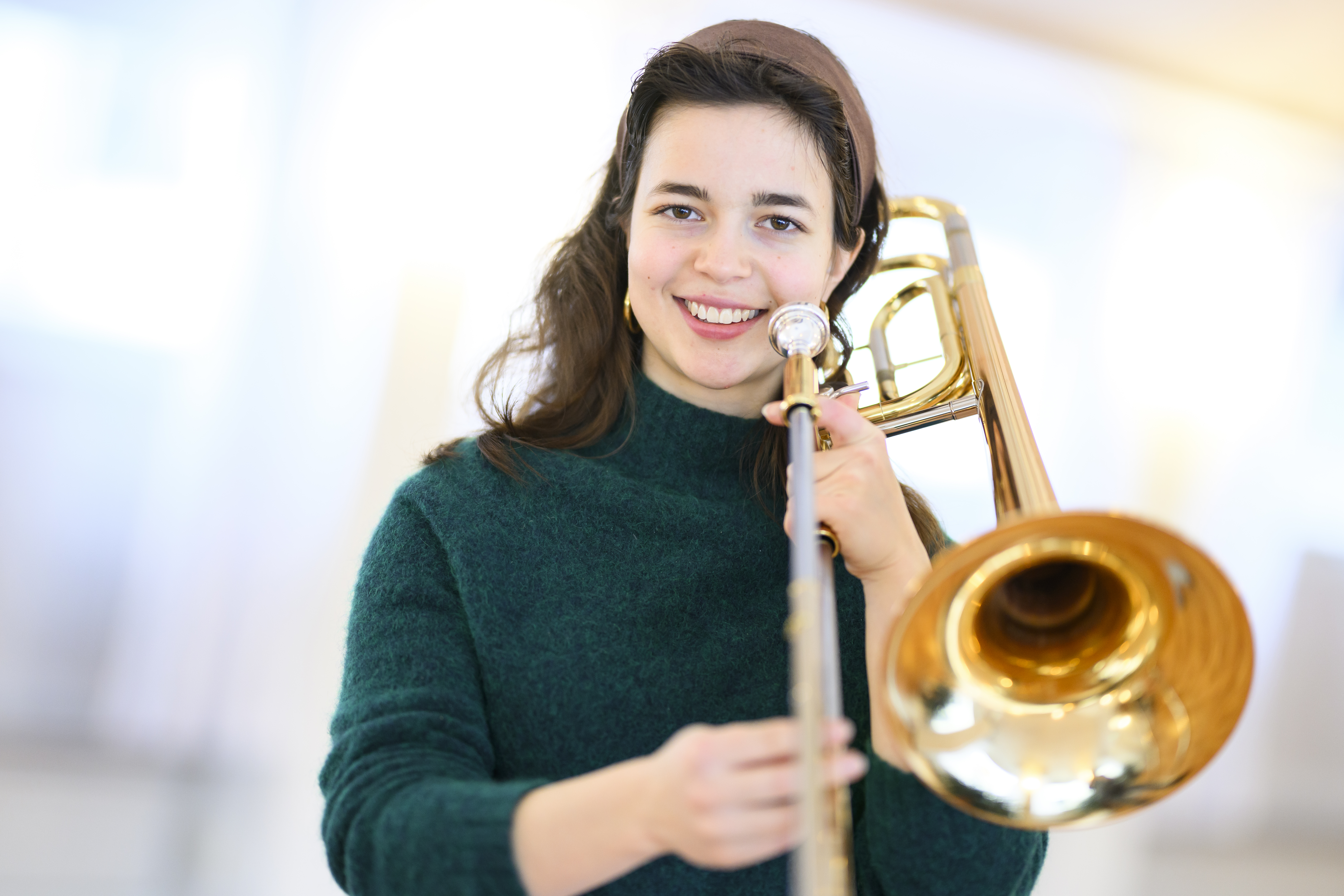 En ung kvinna med brunt hår ler in i kameran. Hon håller i en trombon.