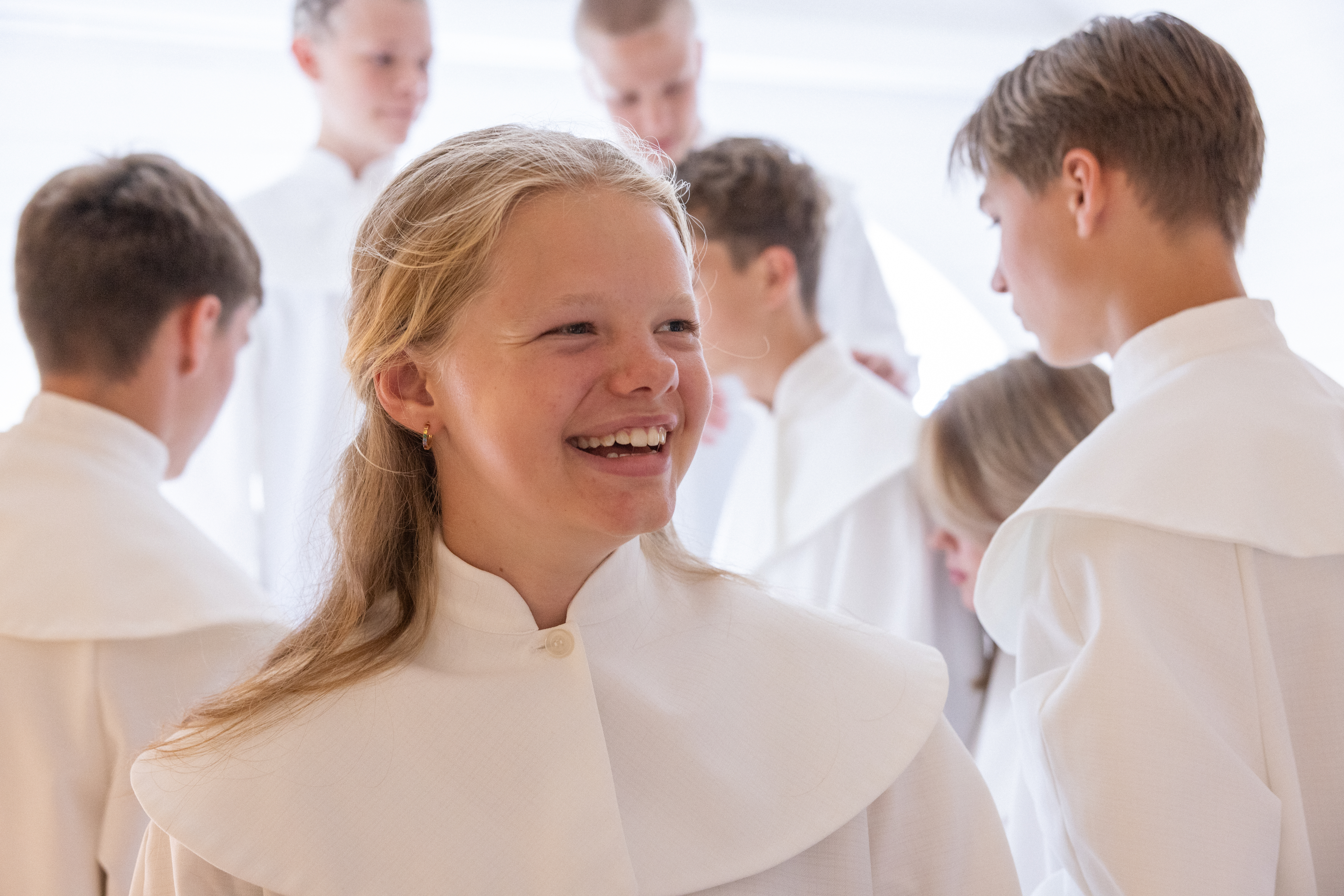 Glad tjej i konfirmandkåpa och andra konfirmander i bakgrunden.