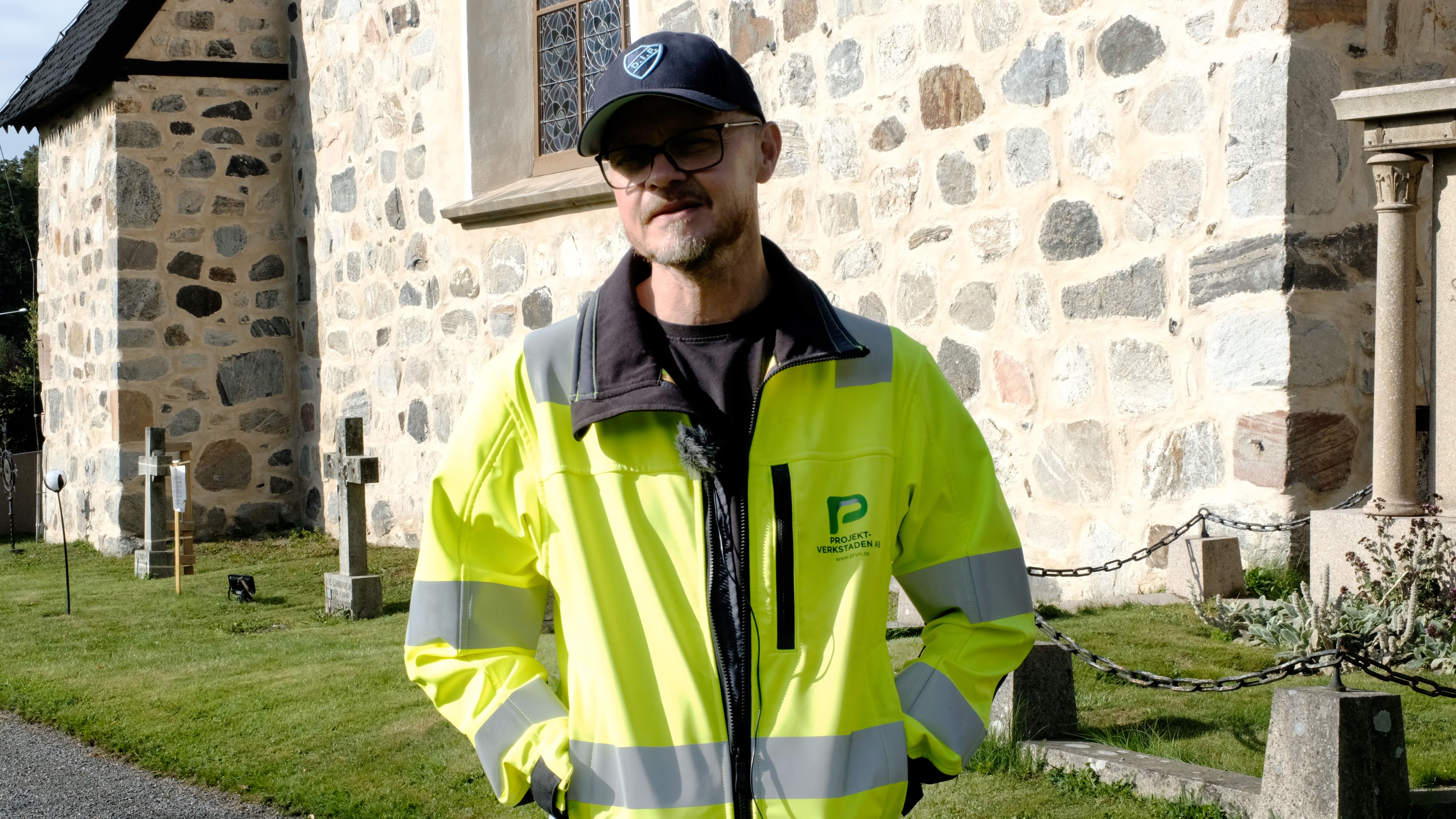 En man i varseljacka och keps tittar strax till vänster om kameran. Han står utomhus. I bakgrunden syns en ljus stenkyrka, gräs och några gravstenar