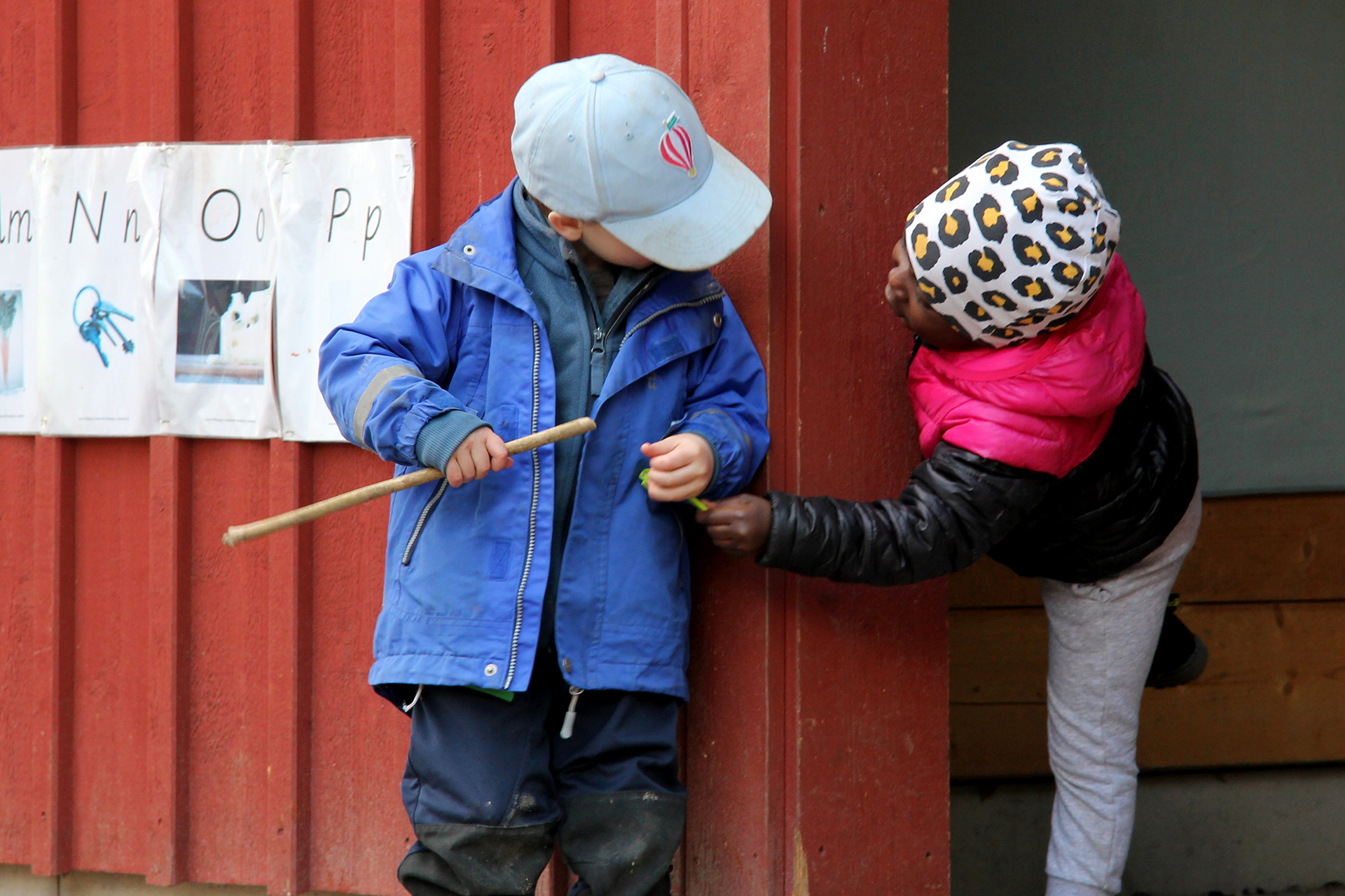 Två små barn i keps och toppluva leker tittut vid en rödmålad husknut på Prästgårdens förskolas utegård.