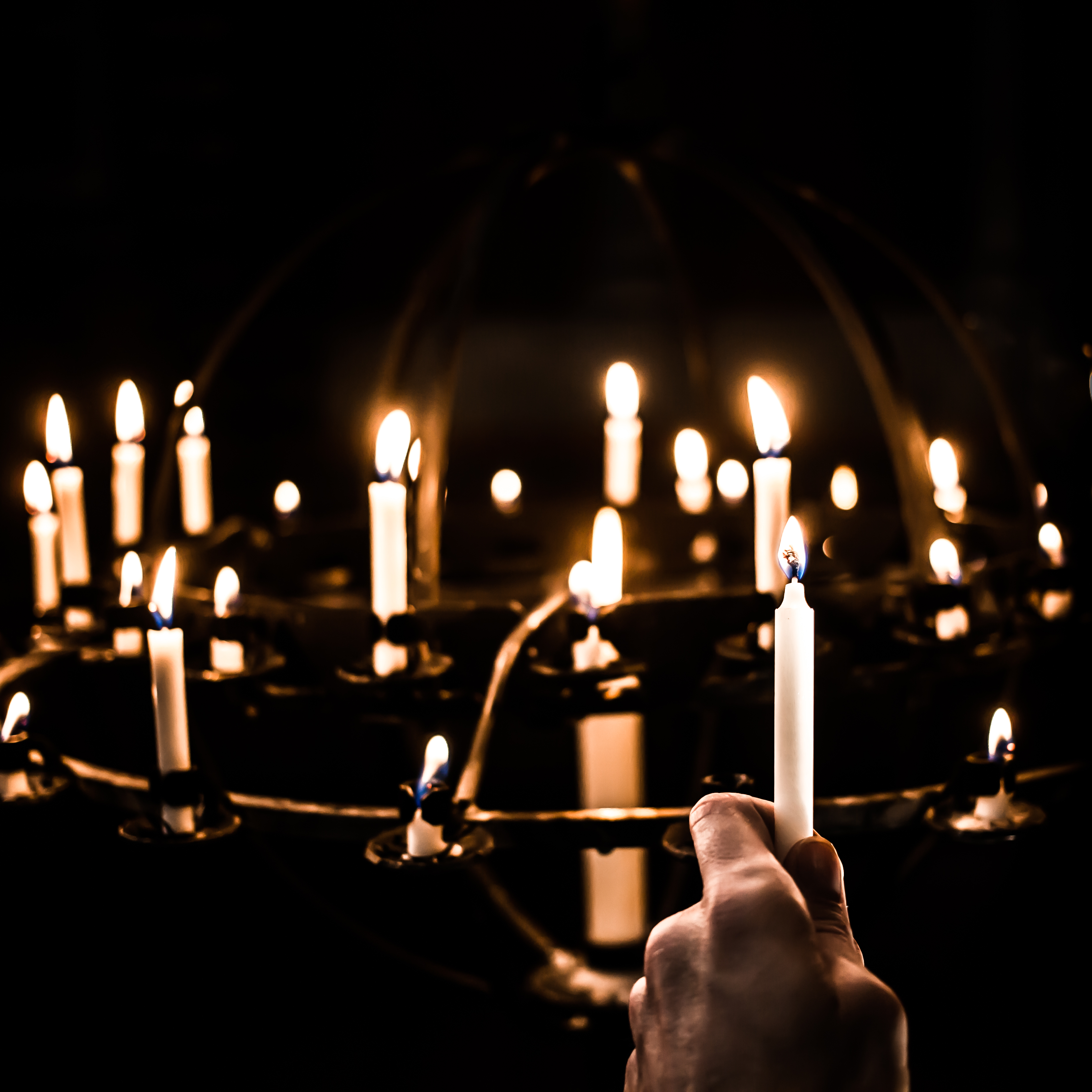 Man ser en ljusbärare full med tända ljus och hand som håller i ett ljus som om man precis tänt det ska sätta det i ljusbäraren.