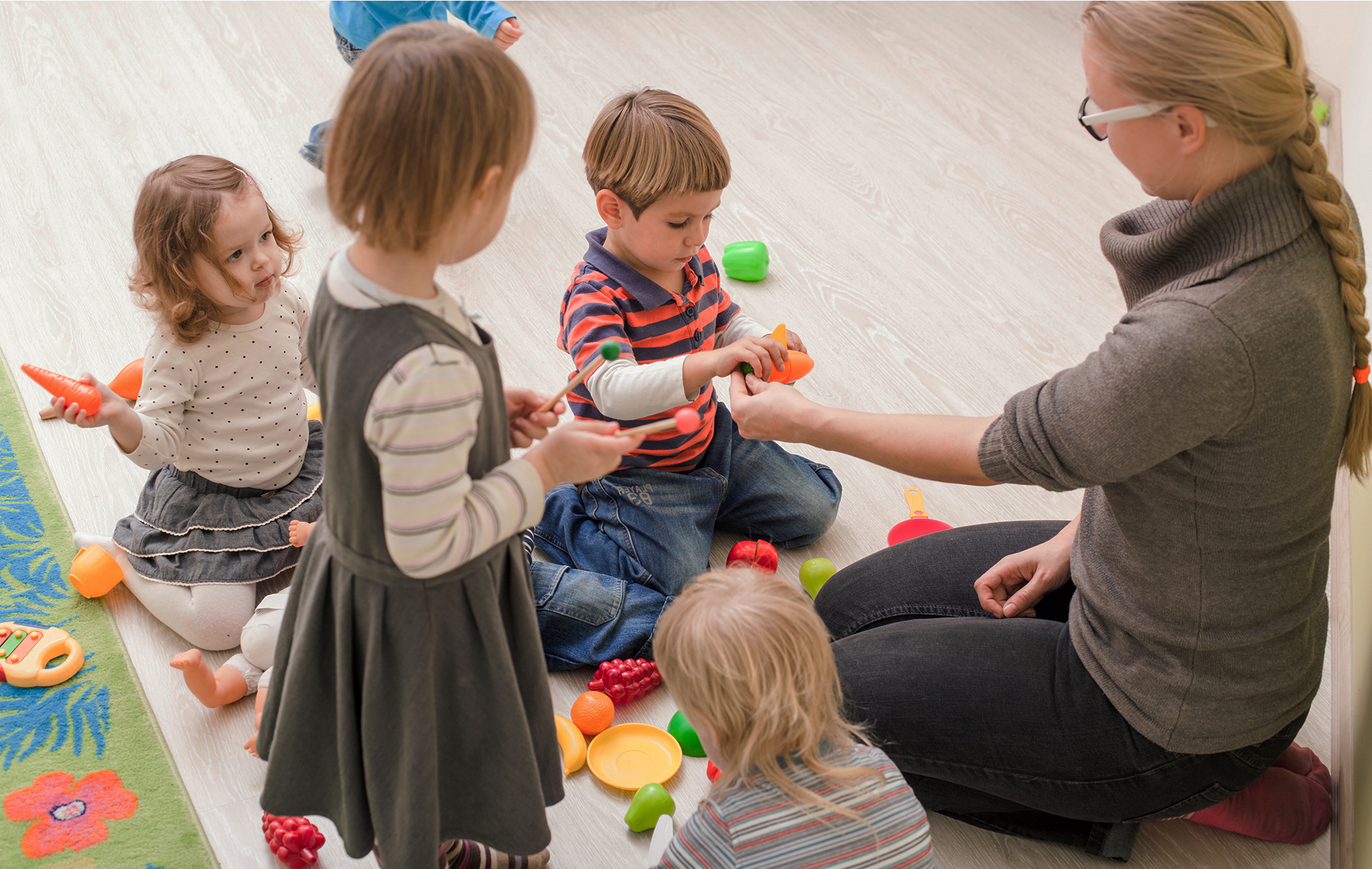 Några barn sitter och står i närheten av en vuxen person som sitter med knäna på golvet.
