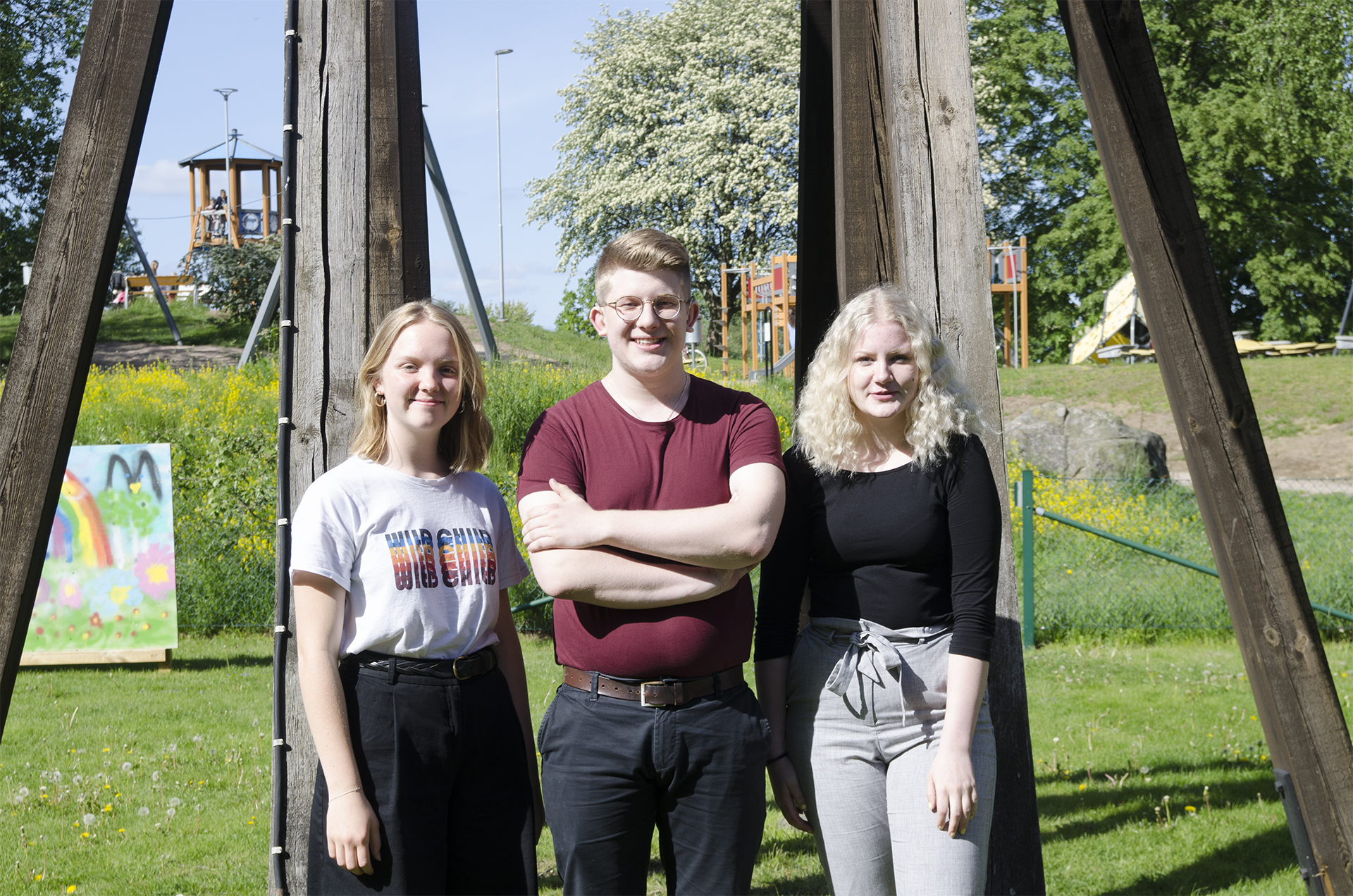 De tre ungdomarna Stina, Hugo och Hanna står utomhus framför klockstapeln i Älmhults församling