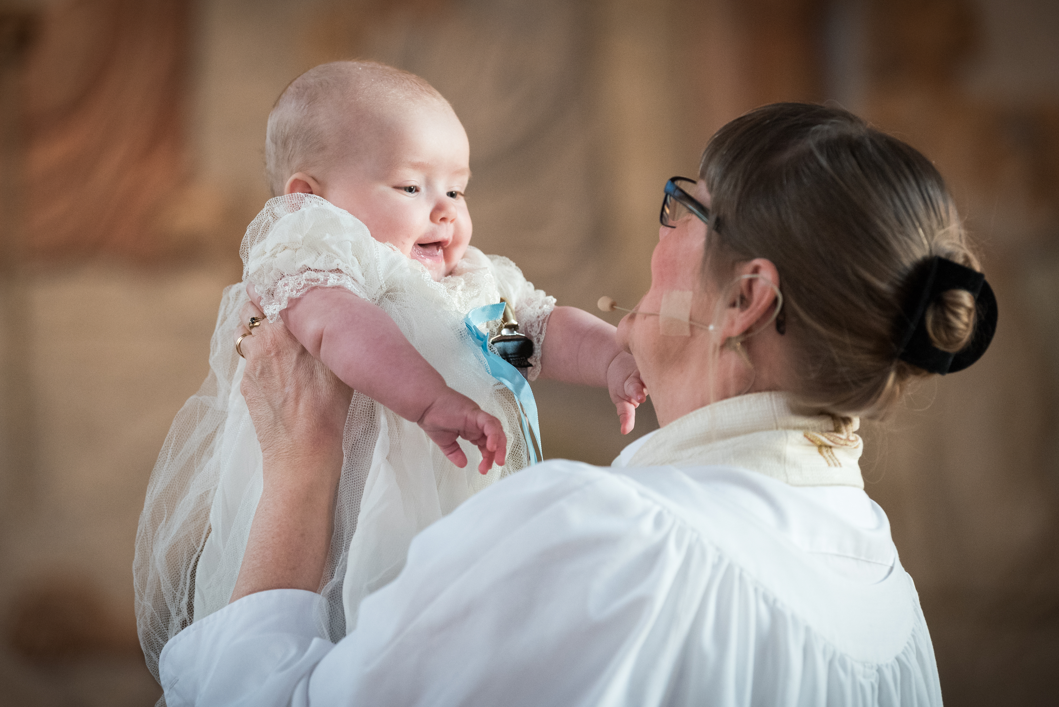 Präst som håller upp en leende bebis i dopklänning.