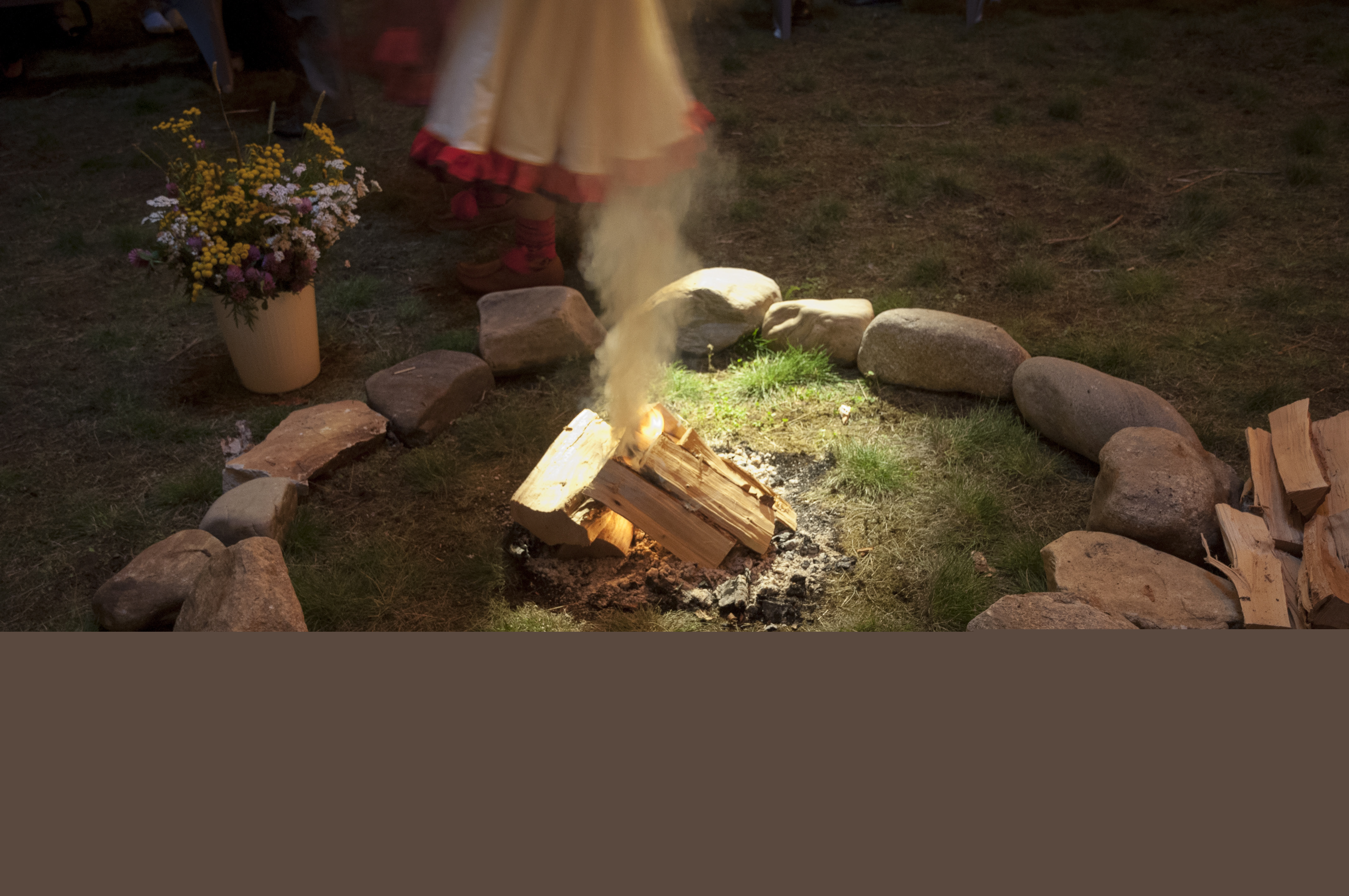 Någon med en samisk klänning och samiska skor står vid en eldstad med liten brasa och en hink med sommarblommor.