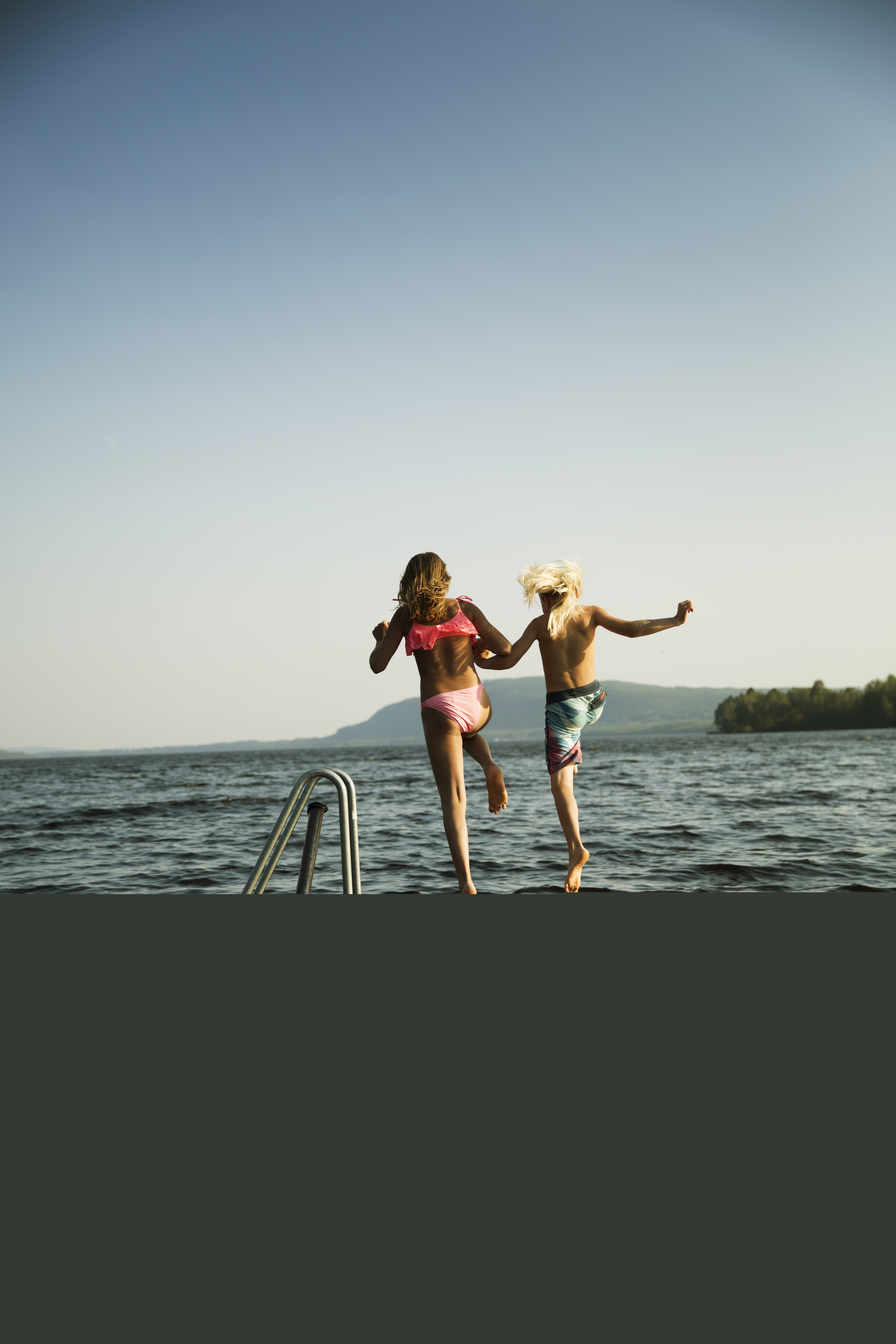 Två barn håller hand och hoppar från bryggan i vattnet.