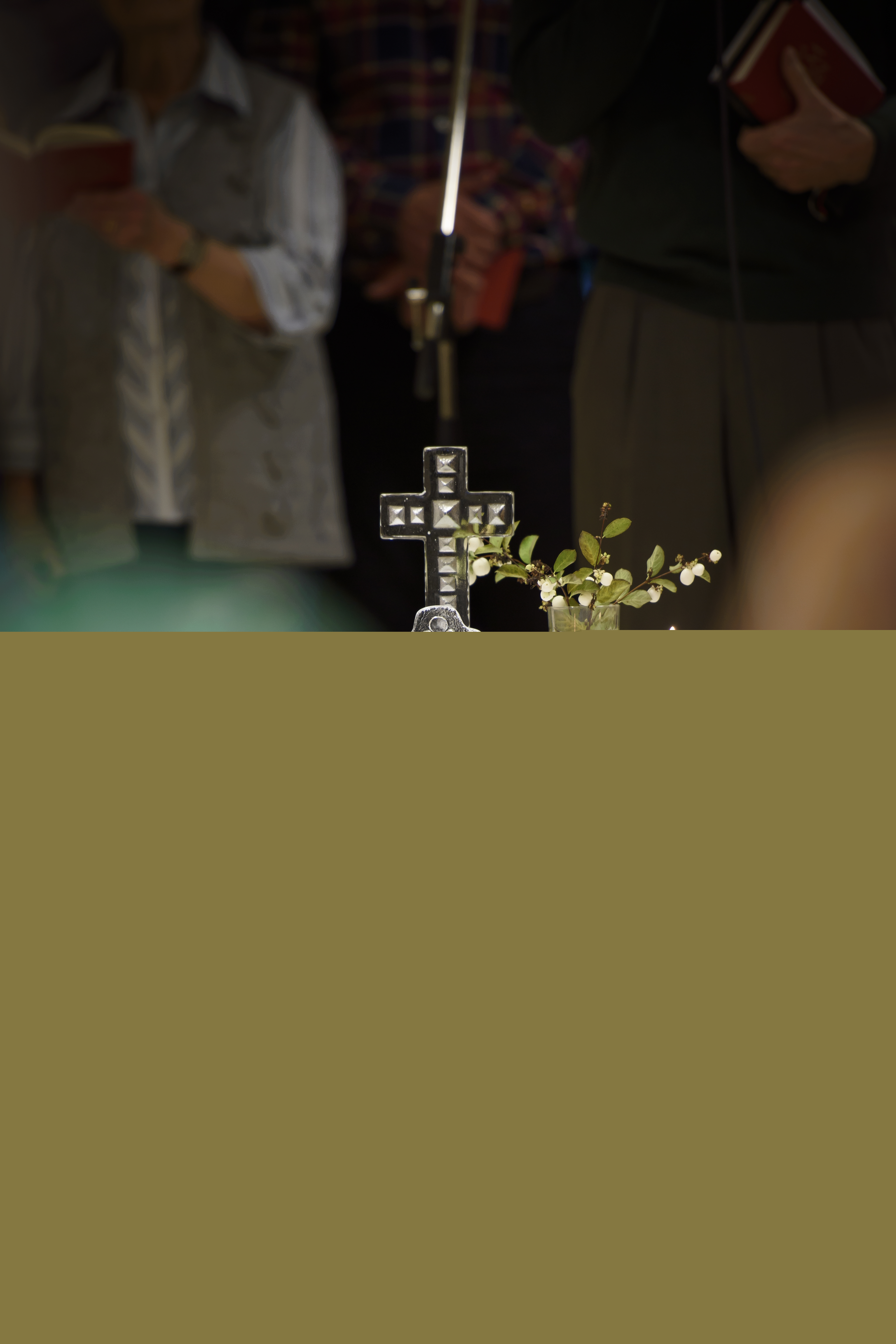 Ett kors, en växt och tända ljus står på ett bord med vit duk. Några suddiga personer står runt om.