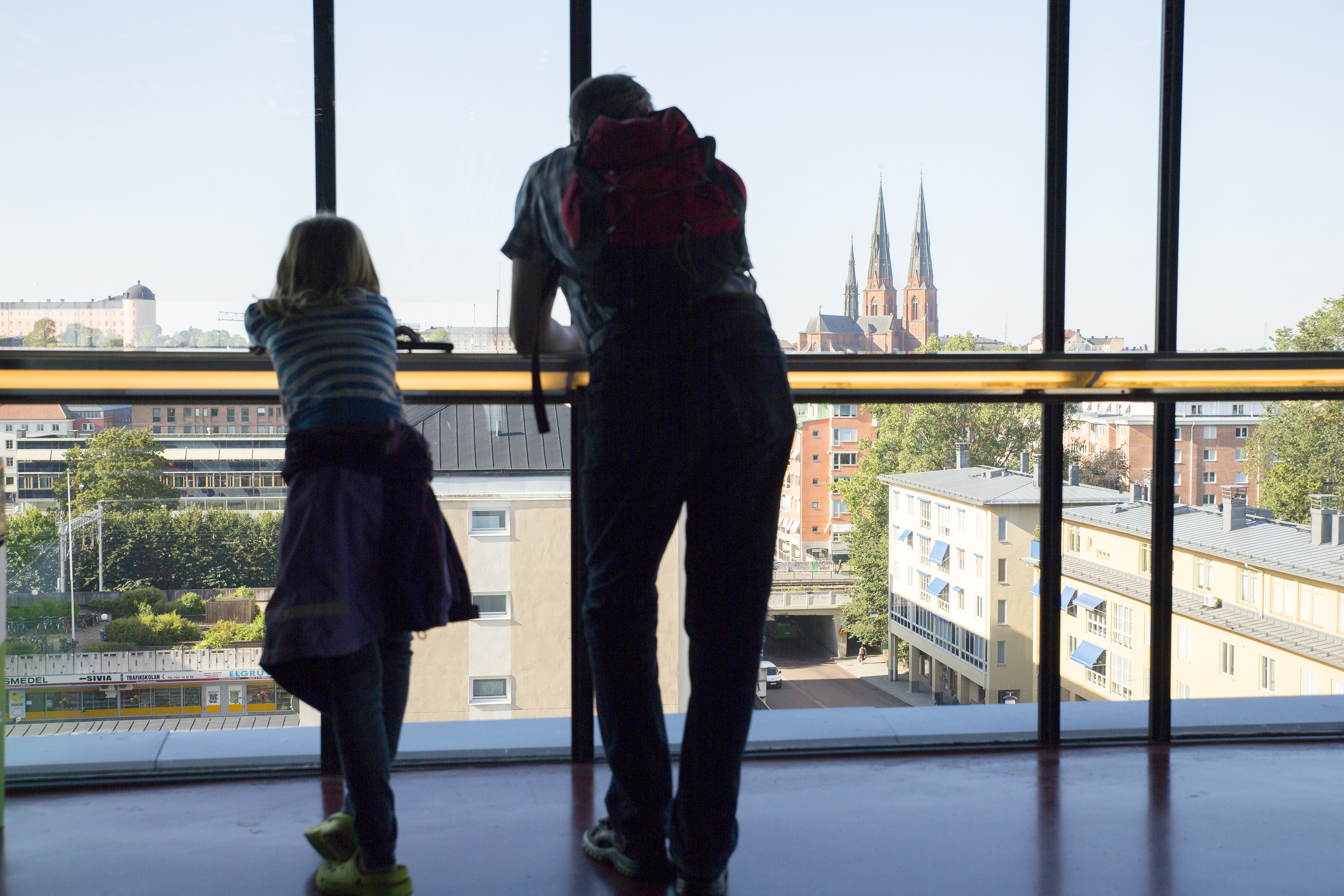En pappa och dotter står i ett höghus och tittar ut genom en glasvägg. Uppsala slott och domkyrkan syns i bakgrunden.