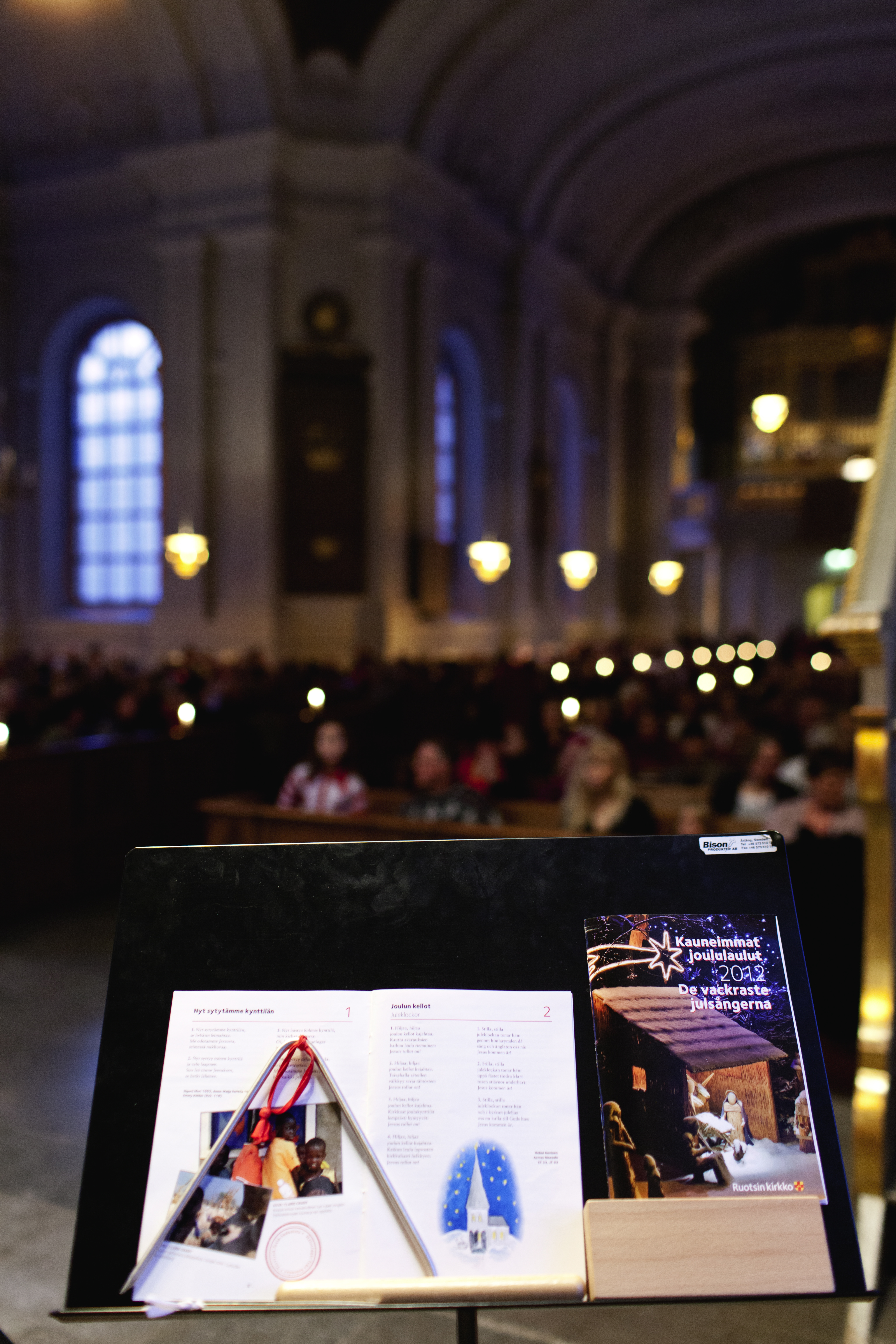 Ett sånghäfte och en triangel ligger på ett notställ. Fullsatta kyrkbänkar i bakgrunden.