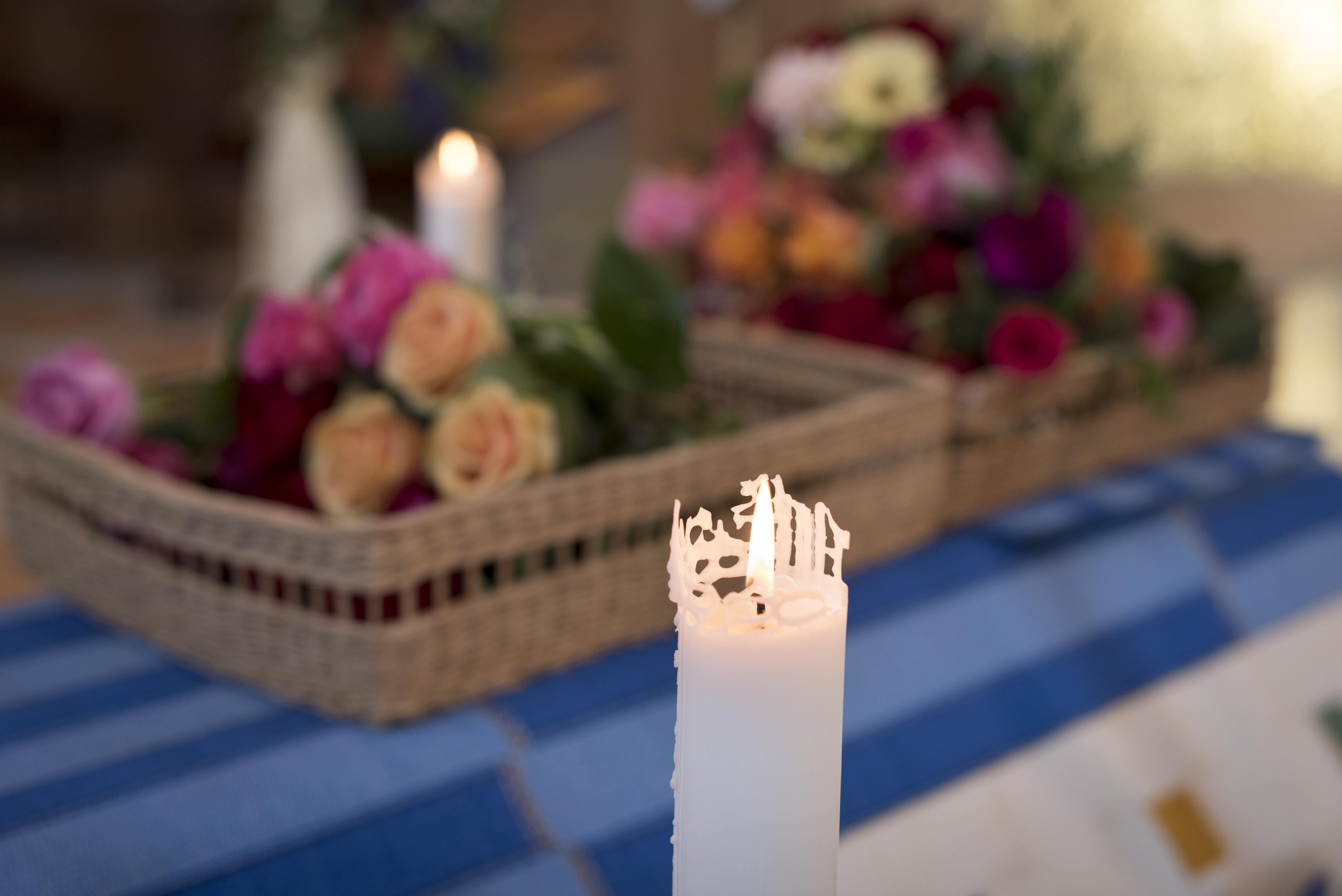 Ett tänt stearinljus. I bakgrunden syns korgar med blommor på en kista.