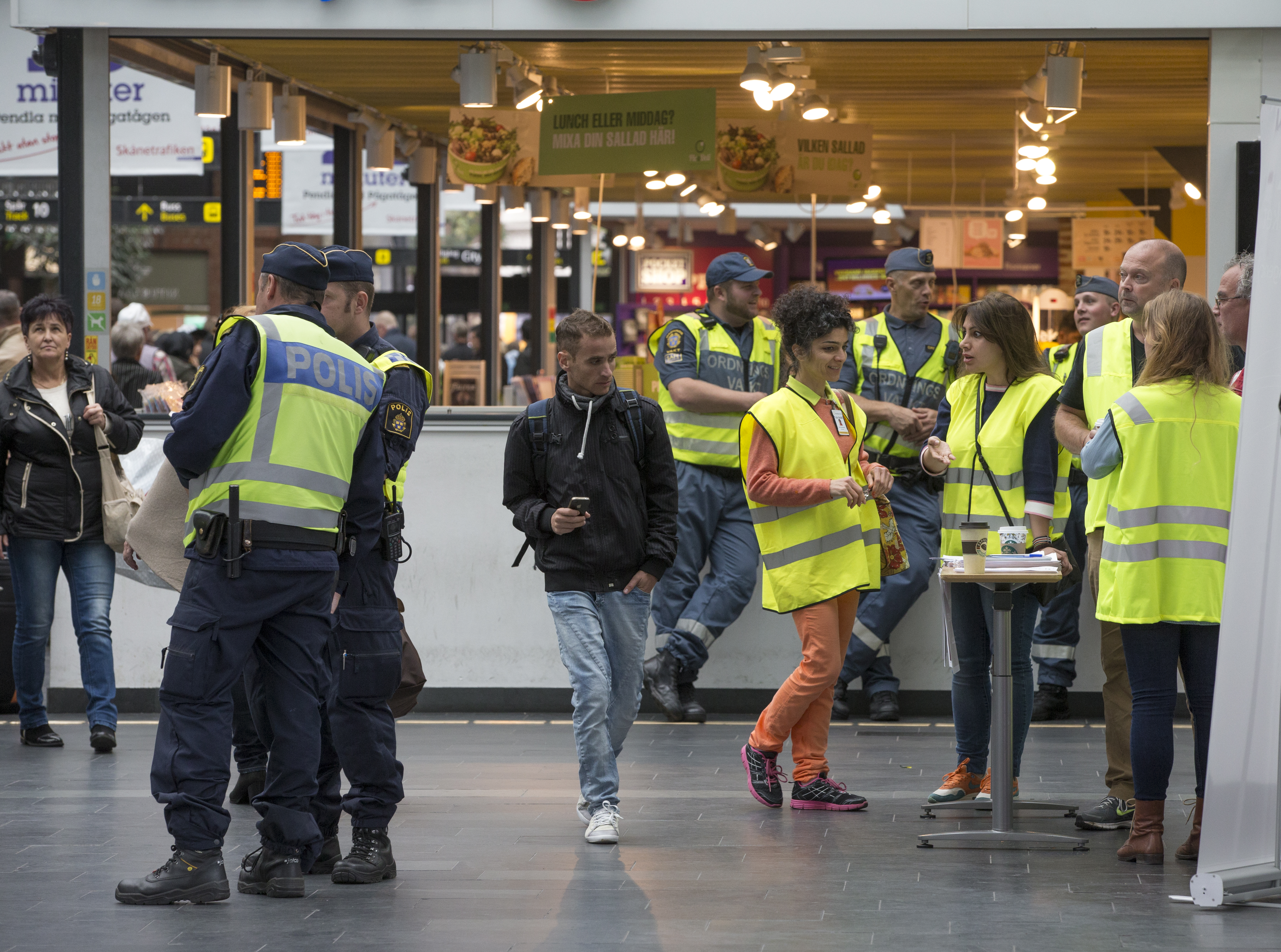 Några ordningsvakter, två poliser och några personer i gula reflexvästar står inne på en galleria.