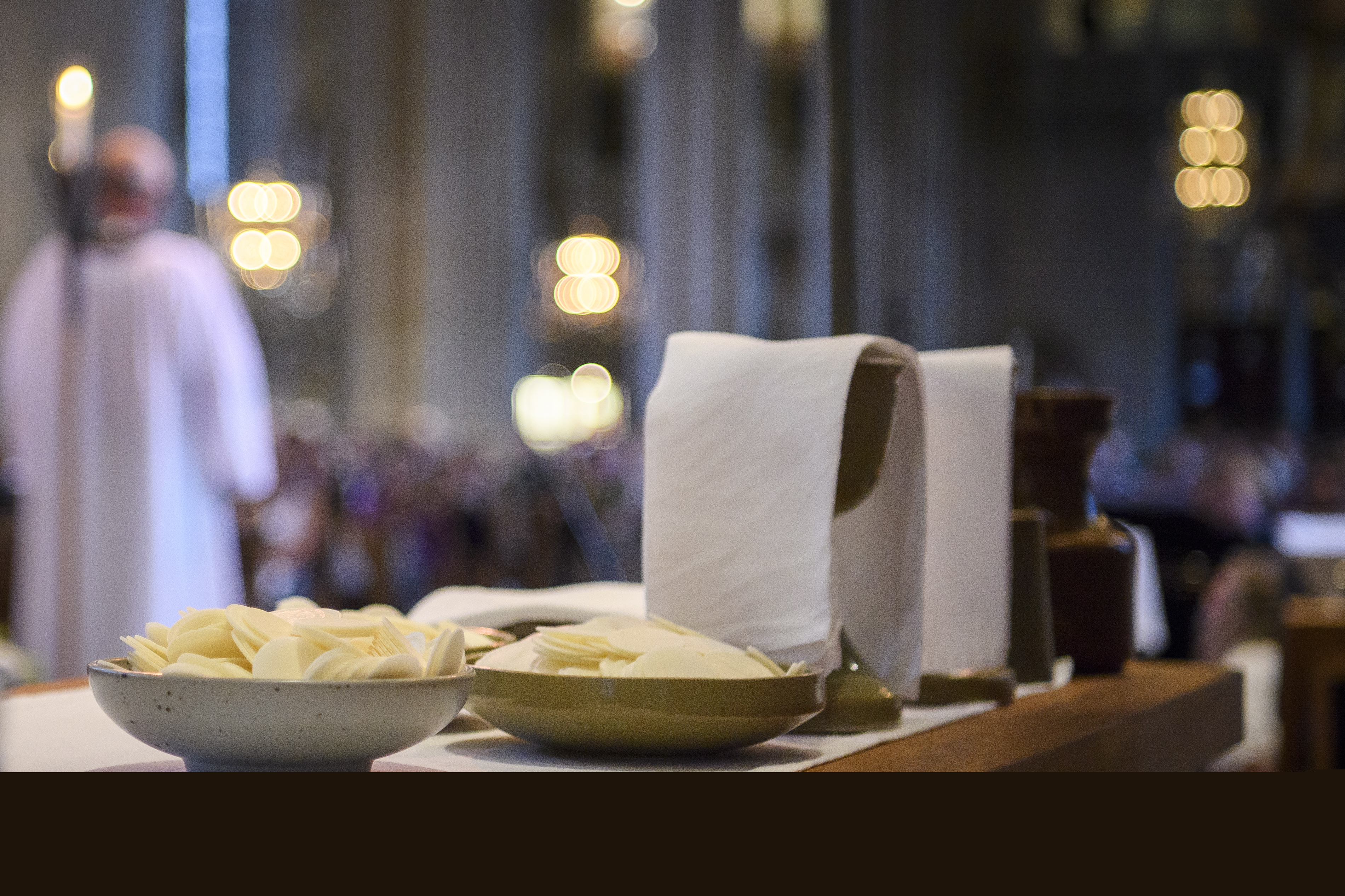Nattvardsvin och oblat står uppdukat på ett bord. En präst och en många kyrkobesökare skymtas i bakgrunden.