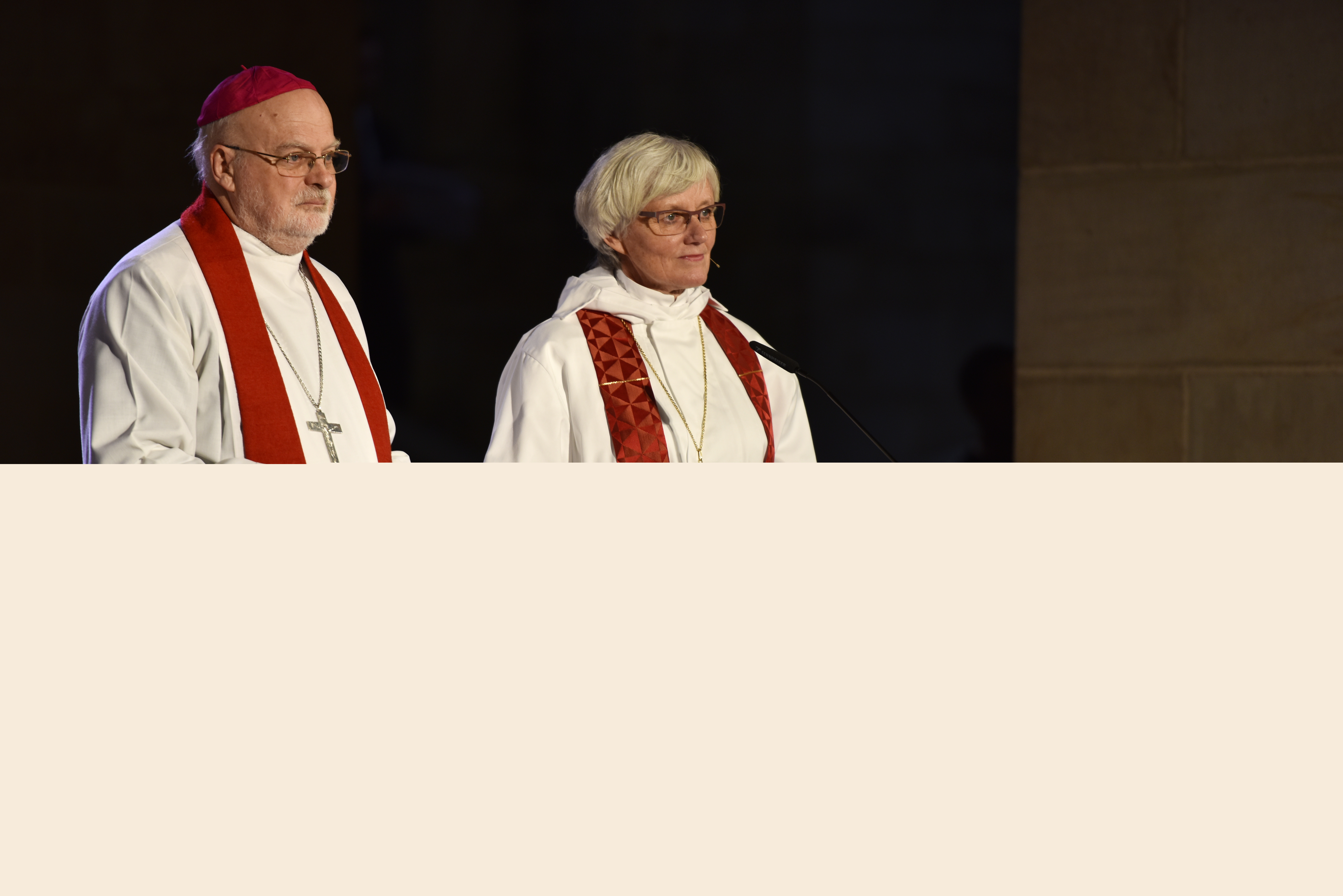 Biskoparna Anders Arborelius och Antje Jackelén står i strålkastarljus i en kyrka och talar i en mikrofon.