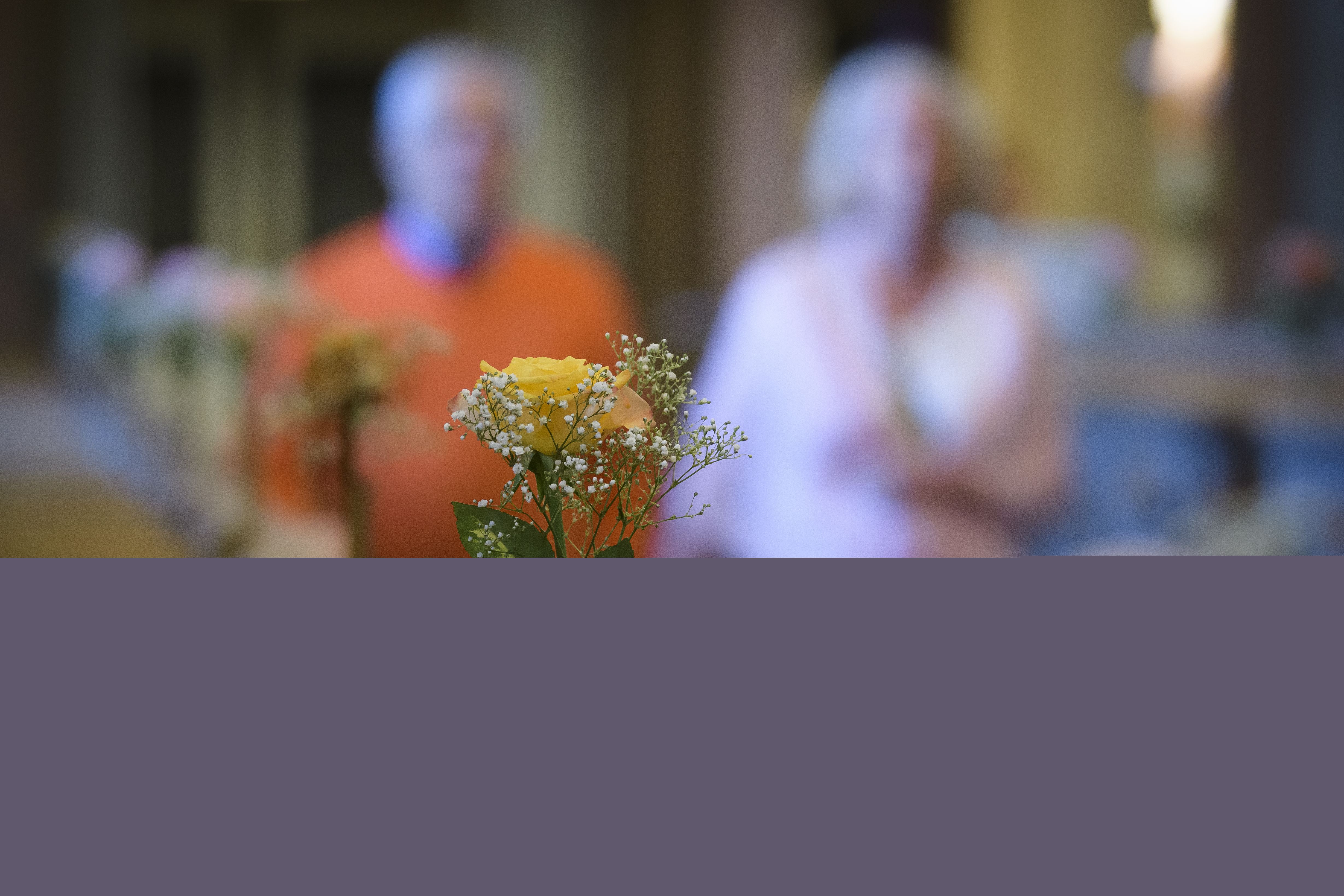 Små vaser med rosor står på bänkraderna i en kyrka. En man och en kvinna syns suddigt gå hand i hand i mittgången.